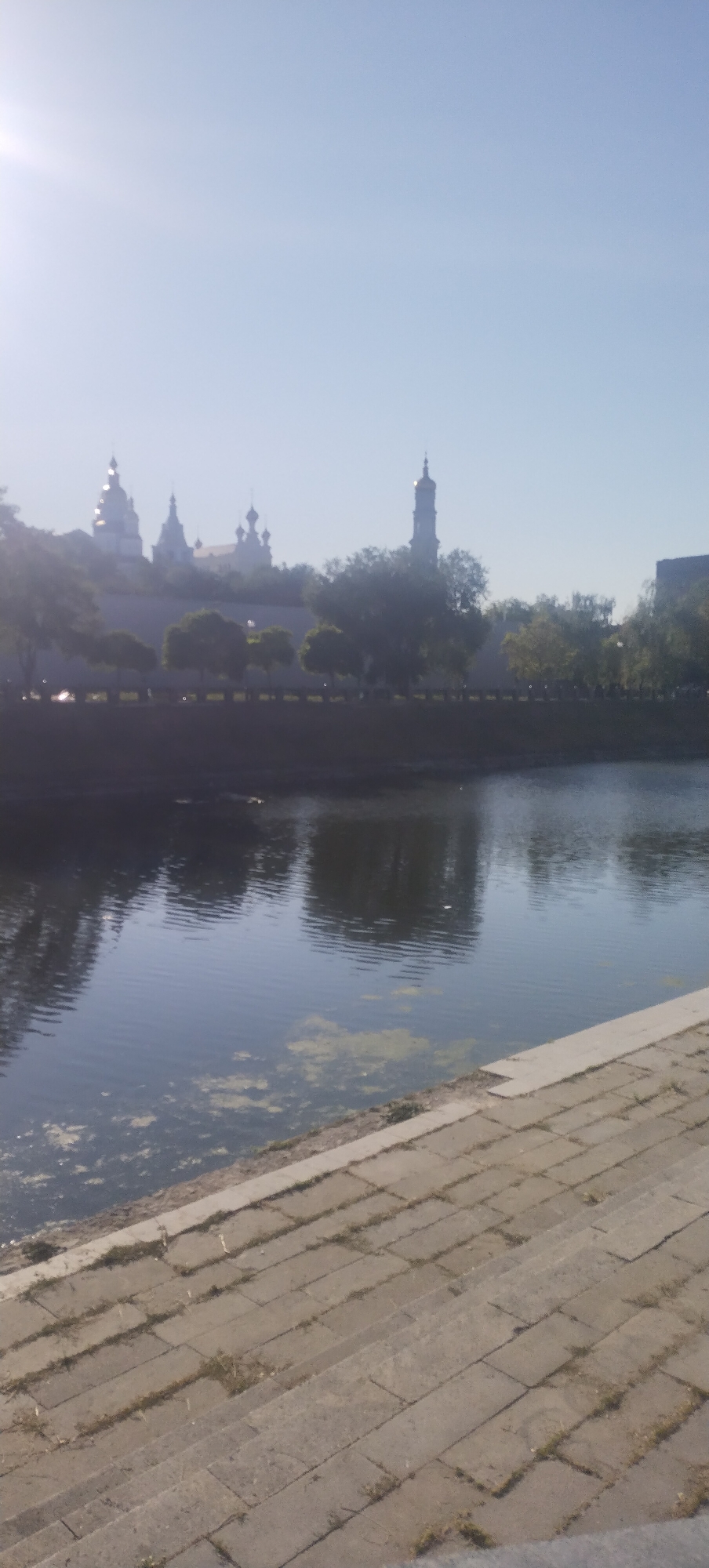 A wide-angle, vertical, medium-shot captures a serene river scene in front of a city skyline under a hazy, sunlit sky. In the foreground, stone steps descend towards the river, leading the eye to the calm, dark water that reflects the sky and the distant structures. Patches of green algae float on the surface. Across the river, a stone embankment is lined with trees, behind which a collection of domed buildings, likely churches or monasteries, are visible. A tall, slender tower stands out amongst them. The buildings and trees are slightly blurred due to the distance and the lighting conditions. The overall atmosphere is peaceful and somewhat ethereal due to the soft light.