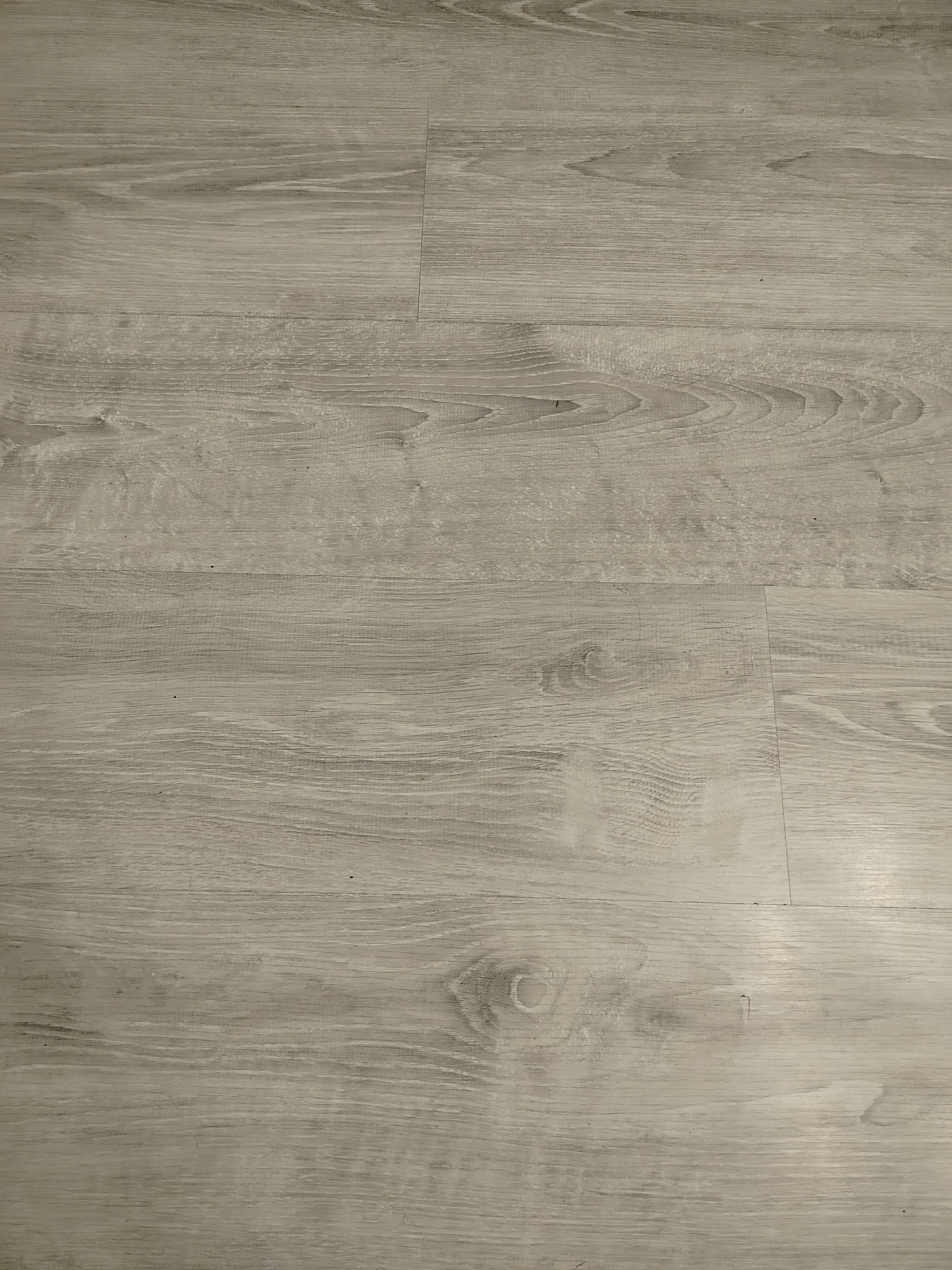 The image shows a close-up of light-colored wood-look flooring, likely laminate or vinyl. The planks run horizontally and are clearly delineated with seams. The wood grain pattern is visible, suggesting a light grey or beige tone. No people, objects, or additional context are present in the image. The location is not discernible from the provided image.