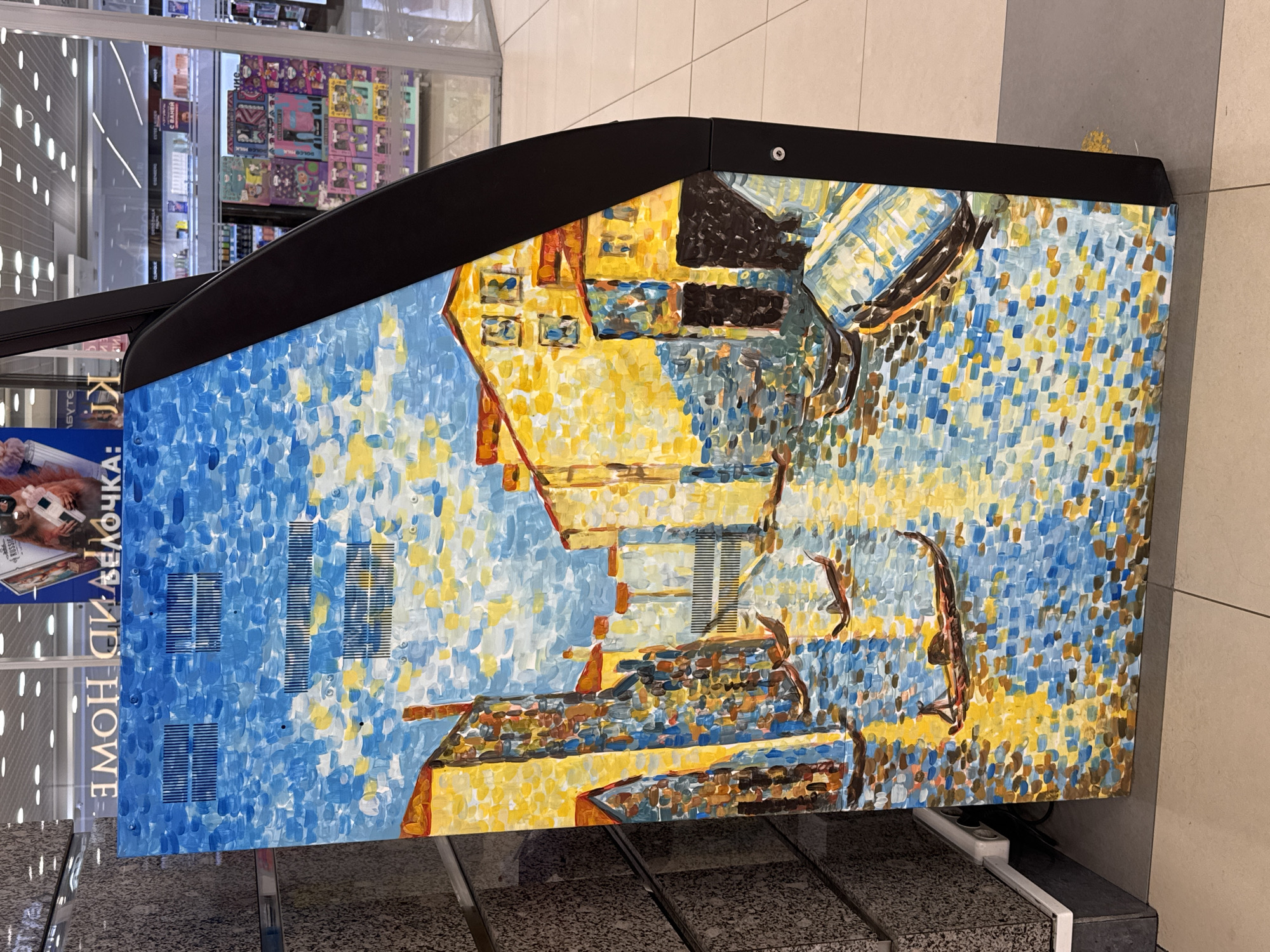 The image shows a commercial display in what appears to be a retail setting, based on the items visible in the background. The display features a painting in a pointillist style, predominantly in shades of blue, yellow, and brown. The painting depicts a building and figures. The Russian text "ИДЕЯ ВЕЛОЧКА: ДОМ" is visible on the left side of the display. The display is located in the city of Saint Petersburg, Russia, as indicated by the Russian text. The flooring is composed of neutral-colored tiles. There is no information about the presence of people, subjects, actions, or interactions in the frame.