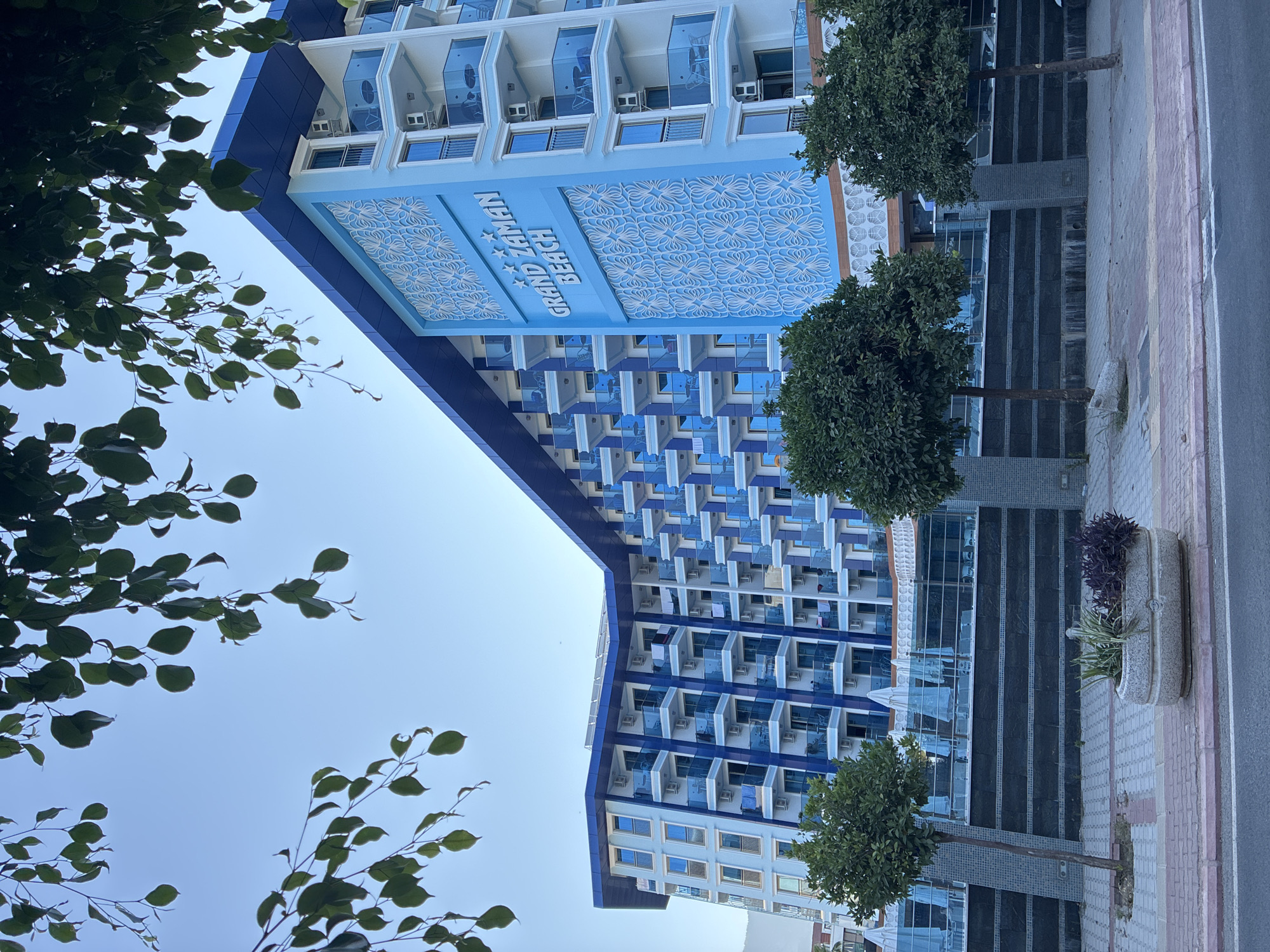 This image depicts the "Grand Zaman Beach" hotel in Alanya, Turkey, viewed from across a street. The hotel is a multi-story building with a striking blue and white facade. It features numerous balconies with blue glass railings and white air conditioning units visible. The hotel's name is prominently displayed in large, white letters on a light blue section of the building, accompanied by five stars. Decorative white patterns adorn this section of the facade.
In the foreground, a sidewalk is visible, paved with interlocking stones in shades of red and grey. A large, oval stone planter sits on the sidewalk, containing green and dark purple plants. Two small, well-maintained trees with full green foliage are planted on the sidewalk, one to the left and one to the right of the planter. Behind the planter and trees, a dark, textured wall forms a boundary, possibly for the hotel's grounds. This wall is topped with a black fence and white umbrellas are visible behind it, suggesting an outdoor seating or pool area.
The background shows a clear blue sky, with the upper branches of a leafy tree obscuring parts of the sky and the top of the hotel. The architecture of the hotel is modern and appears to be designed for a resort or holiday destination, consistent with its location in Alanya, a popular tourist city on the Mediterranean coast of Turkey. The overall scene is bright and sunny, indicating daytime. There are no people or vehicles clearly visible in the frame.