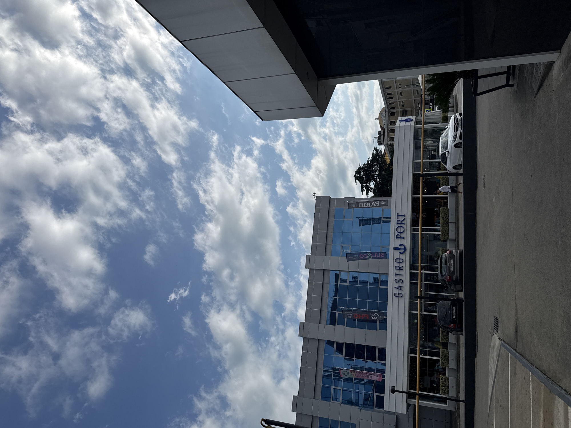 The image, taken in Sochi, Russia, depicts an exterior view of a modern building with large glass windows. The sky above is a clear blue with scattered white clouds, suggesting a bright, daytime setting. The building features the prominent text "GASTRO PORT" in large blue letters, along with a stylized anchor logo.
In front of the building, a white Mercedes-Benz car is parked. Further down the street, another dark-colored car is visible. A person in a white t-shirt and dark shorts is walking on the sidewalk. The architecture of the building is contemporary, with a sleek facade and multiple levels. Reflections of the sky and clouds are visible on the glass surfaces of the building.
Beyond the modern structure, older buildings with classical architectural elements can be seen in the background, including a domed structure. Trees, including a mature evergreen, are also present, adding to the urban landscape. The overall scene conveys a sense of a pleasant day in a city that blends modern development with older architecture. The presence of the "GASTRO PORT" sign indicates a commercial establishment, likely a restaurant or a complex housing food-related businesses. The image is oriented vertically, with the top of the frame showing the sky and the lower part showing the ground and the building.