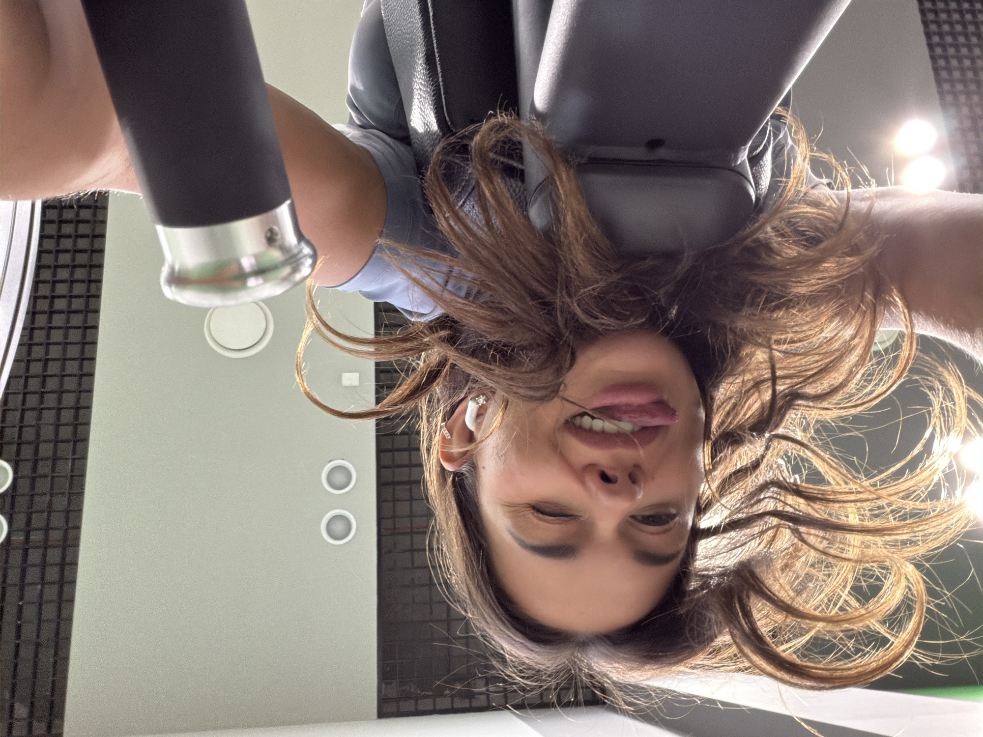 A woman with shoulder-length brown hair, wearing an earbud in her left ear and a light blue shirt, is upside down and sticking her tongue out. She is winking her right eye. She is likely at a gym or fitness center as there are exercise machine components directly above her and light fixtures on the wall. The specific location in Krasnoyarsk, Russia is not discernable from the image.