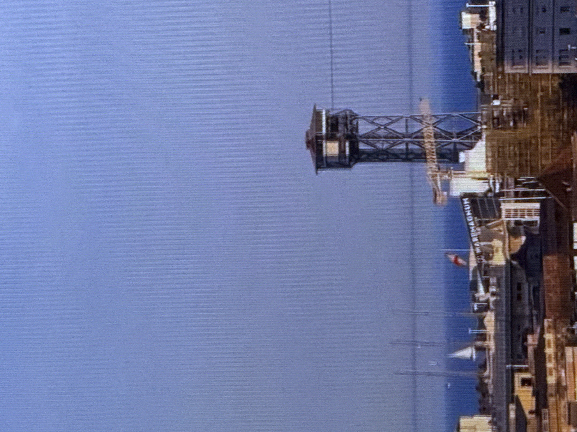 This image, taken in Dubai, United Arab Emirates, depicts a waterfront scene with industrial elements and a clear blue sky. The primary focus is a tall, metallic tower structure, possibly a crane or part of a construction, dominating the left side of the frame. Its intricate latticework is clearly visible against the flat expanse of blue water.
To the right, a ship or large vessel is docked, displaying a multi-story structure with visible windows and architectural details. A red and white flag is seen flying from one of the ship's masts. Below the ship, a sign reads "MAGNUM." Several smaller masts and structures, potentially part of other boats or harbor infrastructure, are also visible in the water.
The overall impression is one of a bustling port or industrial area. The lighting suggests it is daytime, with the sun casting shadows and illuminating the structures. The water appears calm, indicating fair weather conditions. The image is oriented vertically, with the sky taking up the majority of the left side and the industrial elements and water filling the right. The background is a soft, textured blue, possibly indicating a screen or a digital display.