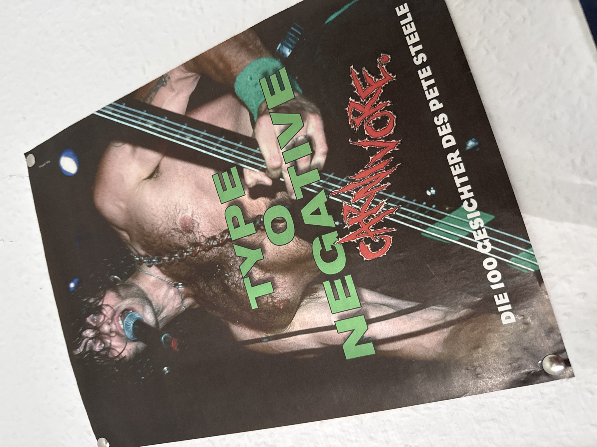 The image shows a close-up of a poster featuring the band Type O Negative. The poster is dark, with the band members illuminated. In the foreground, a man with dark, messy hair is singing into a microphone. He has a chain around his neck and is partially shirtless, revealing a hairy chest. To the right, another band member, shirtless, is playing a guitar, his body partially obscured by the instrument's strings. The band's name, "TYPE O NEGATIVE," is written in large green block letters across the poster, with "CARNIVORE" in a red, stylized font below it. At the bottom, white text reads "DIE 100 GESICHTER DES PETE STEELE." The poster appears to be affixed to a wall, with metal thumbtacks visible in the corners. The background is dark and out of focus, with some blurred lights. There is no indication of the location being in Bad Sachsa, Germany; the image solely depicts the poster itself.