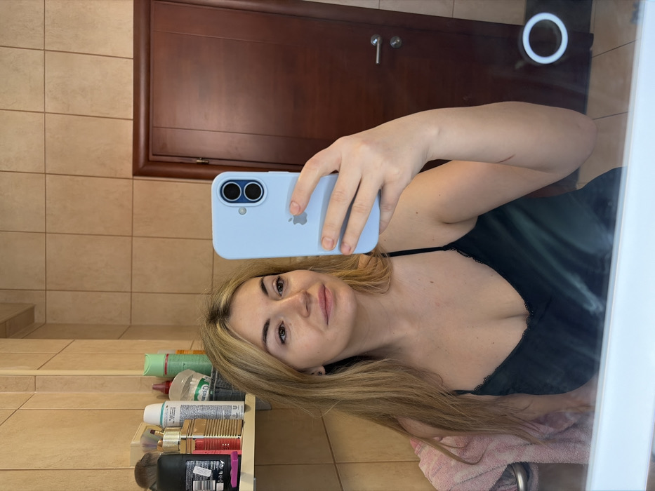The image depicts a mirror selfie taken by a female in a domestic bathroom, located within the city of Budva, Montenegro. The specific street or building details are not ascertainable from the visual evidence.
The central subject is a fair-skinned female with long, straight, blonde hair, wearing a dark, possibly emerald green or black, satin camisole featuring lace trim along the v-neckline. She holds a light blue iPhone, equipped with a dual-lens camera system and an Apple logo on its light blue case, in her raised left hand to capture her reflection. Her gaze is directed slightly off-camera to her right. A reddish linear mark is visible on her right forearm. Her face appears to have minimal or no visible makeup, and a small dark earring is present in her left ear.
The background consists of light brown square wall tiles. To the left, a multi-tiered shelf unit holds various toiletries and cosmetic items, including bottles, brushes, and spray cans. A section of a light-colored towel, potentially pink, is draped over a metallic bar on the lower left. On the right side of the reflection, a dark brown wooden door with a silver handle and keyhole mechanism is visible. A circular illuminated element, consistent with integrated mirror lighting, is reflected on the lower right portion of the mirror surface. The bottom edge of a white countertop or basin is partially visible below the mirror.