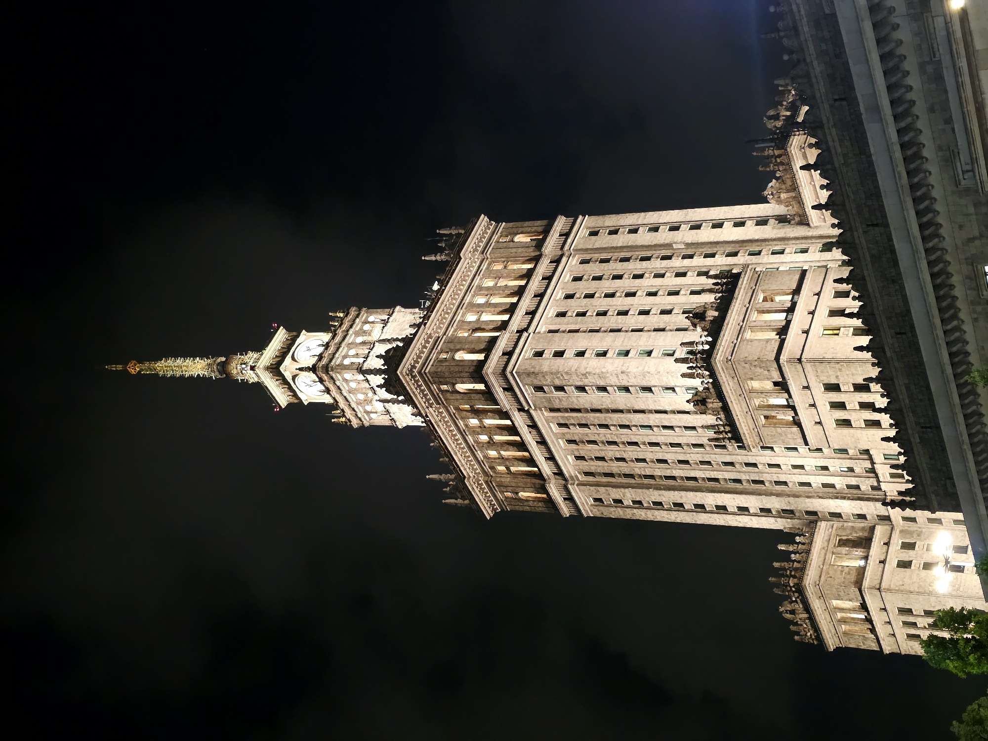 The image shows the Palace of Culture and Science in Warsaw, Poland at night. The skyscraper is illuminated, with lights on in many of its windows. The building's ornate spires and architectural details are visible against the dark, cloudy sky. The foreground shows a portion of another building, likely connected to or in front of the Palace. There are no people visible in the image. The scene is static and depicts the building and its surroundings at night.