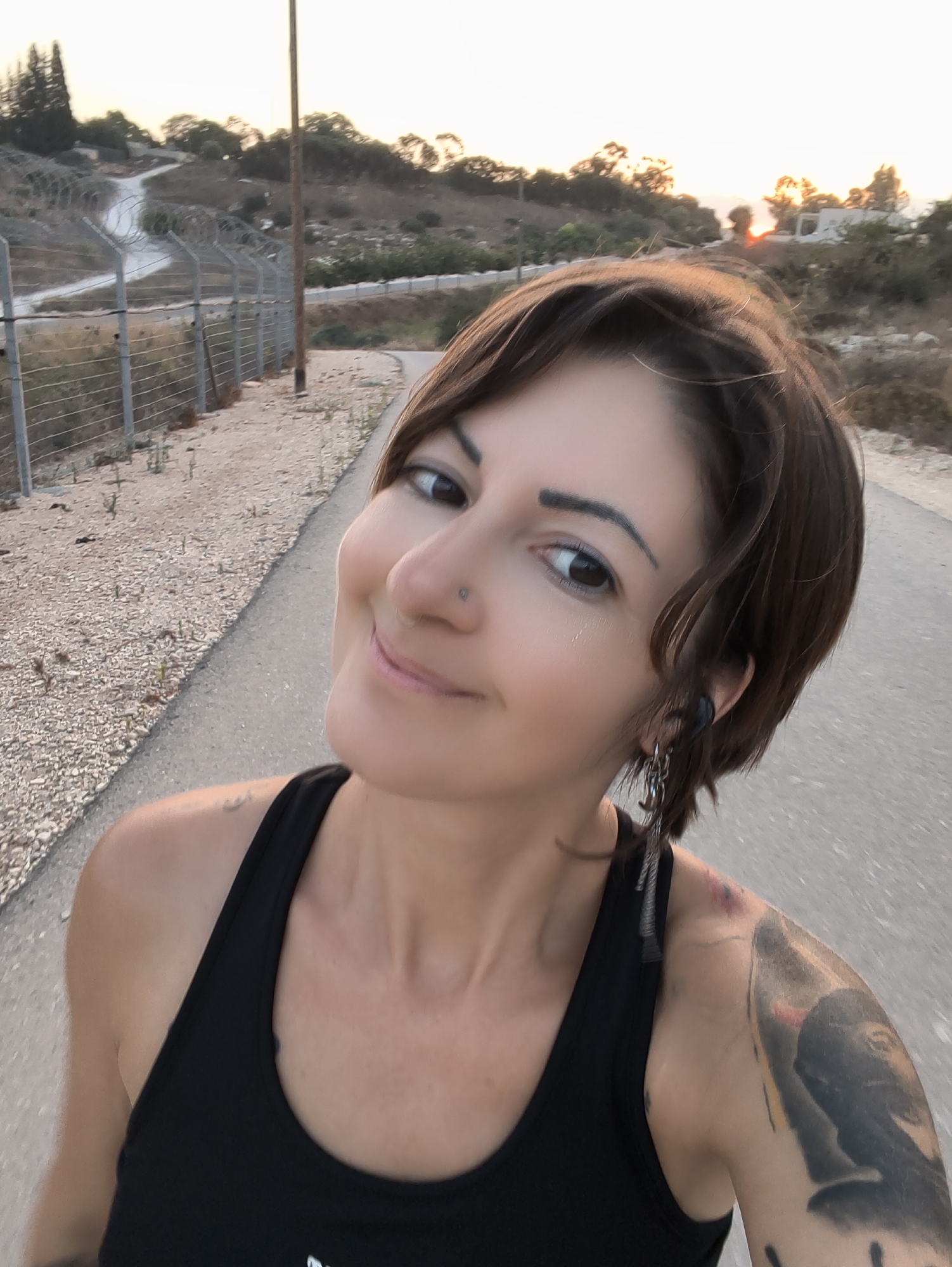 A woman with short brown hair and brown eyes is pictured from the chest up, facing slightly to the left. She is wearing a black tank top and a silver hoop nose ring. Her left ear has a black earbud and a dangling earring with multiple charms. A large tattoo is visible on her right shoulder and upper arm, depicting a person's face. She is outdoors, on a paved road with a gravel shoulder lined with a barbed wire fence. The background shows dry, hilly terrain with sparse vegetation and trees, and a clear sky with a faint sunset glow. A wooden utility pole stands to the left of the woman, near the fence. The location appears to be a rural or semi-rural area, consistent with the Lachish Regional Council in Israel, given the landscape and vegetation.