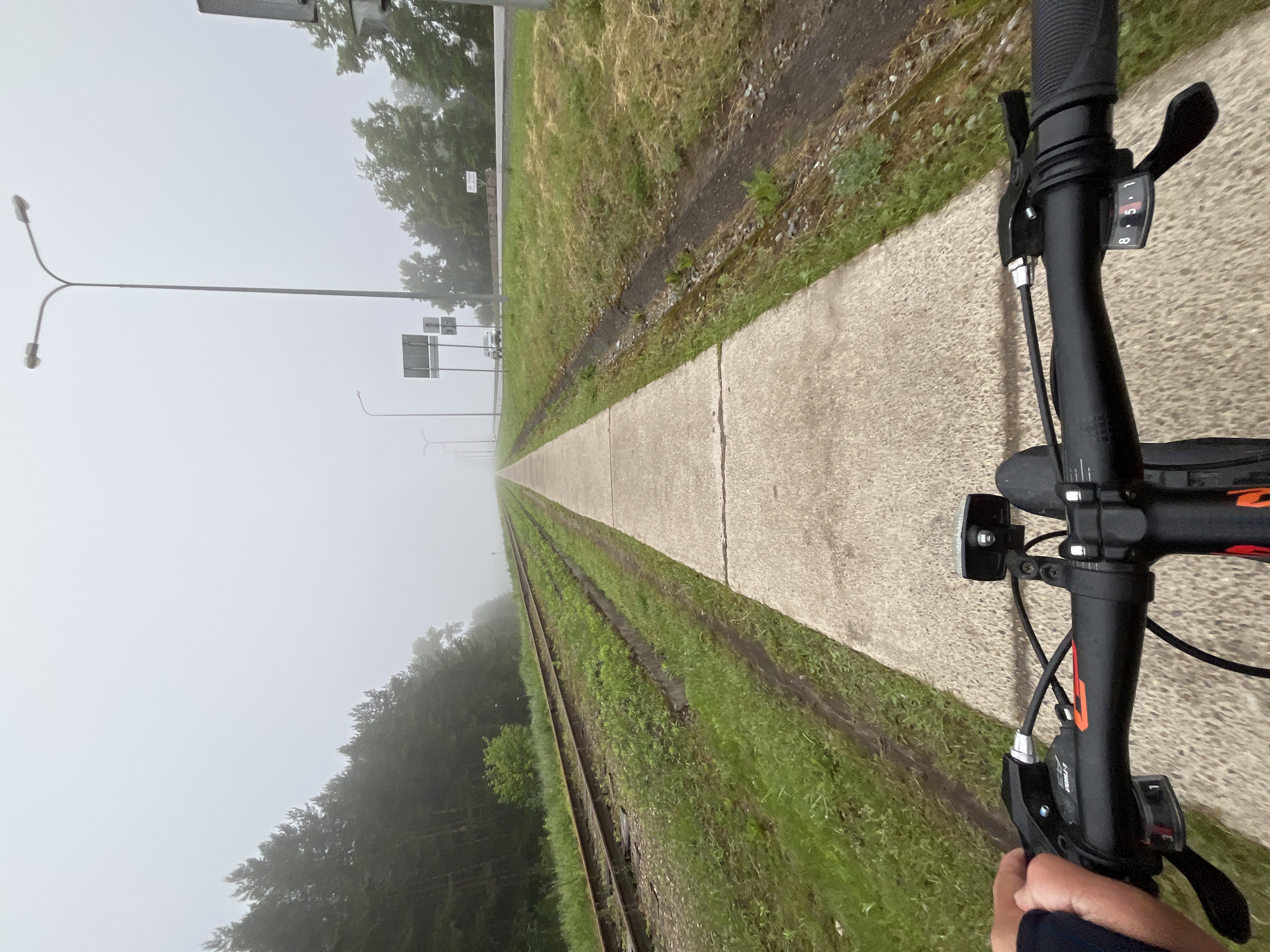 This media file presents a first-person perspective from a bicycle ride along a concrete path, shrouded in thick fog. The viewpoint is from the cyclist's handlebars, which are visible in the foreground on the right side of the frame. A hand grips the right handlebar, where a shifter clearly displays the numbers "8 5 1," indicating the current gear. Another shifter/brake lever on the left handlebar shows partial text, including "SHIMANO" and possibly "ALIVIO." A bicycle front light is also attached to the handlebars.
The concrete pathway extends straight into the hazy distance on the right, bordered on its left by a strip of green grass and dirt. Parallel to the path and grass, a set of railway tracks stretches forward, gradually disappearing into the dense fog. Further to the left, a line of trees appears as blurred, dark masses against the white mist. In the far distance, tall street light poles, including one with a distinctive double-lamp design, and indistinct road signs are barely visible through the heavy fog.
The activity depicted is cycling. The weather is notably very foggy or misty, which diffuses the light and obscures the horizon, making it difficult to ascertain the exact time of day, though it appears to be daytime. The scene evokes a sense of solitude or peaceful journey through an atmospheric landscape in Chaikovsky, Russia. The overall impression is one of moving forward into an uncertain but quiet environment.