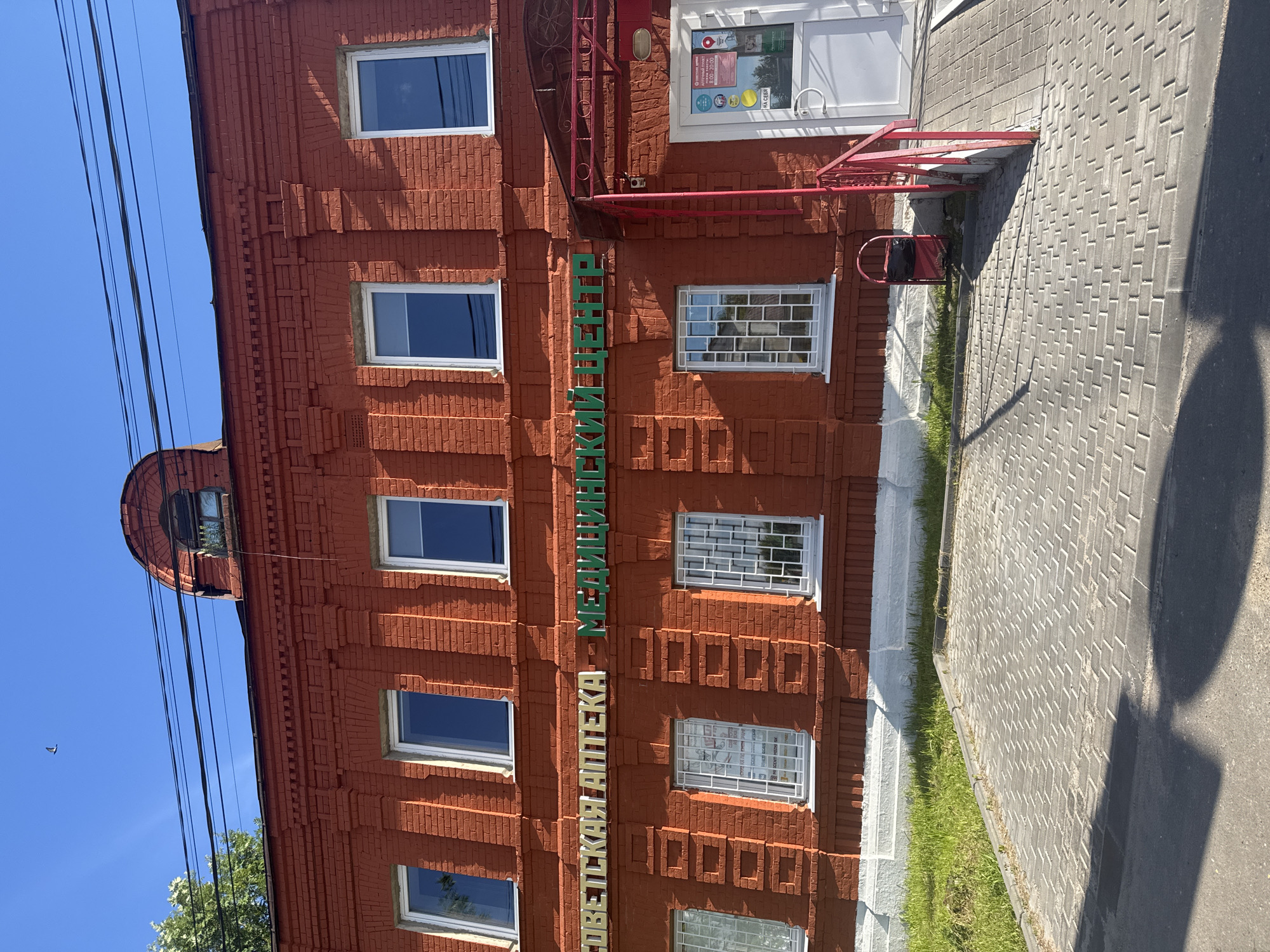In the city of Vereya, Russia, a two-story red brick building with arched windows on the upper floor is shown. The building has several rectangular windows on both floors, some with decorative brickwork above them. A sign in white and green Cyrillic lettering spans the upper facade, reading "СОВЕТСКАЯ АПТЕКА - МЕДИЦИНСКИЙ ЦЕНТР" (Soviet Pharmacy - Medical Center). A red awning extends over the entrance to the building, which is a white door with a glass panel. A ramp leads to the entrance, flanked by a red railing. A red trash receptacle is situated near the entrance. The ground in front of the building is paved with grey bricks, transitioning to asphalt in the foreground. Green grass grows along the base of the building. The sky is clear and blue, with a few dark power lines running across the top of the frame.
