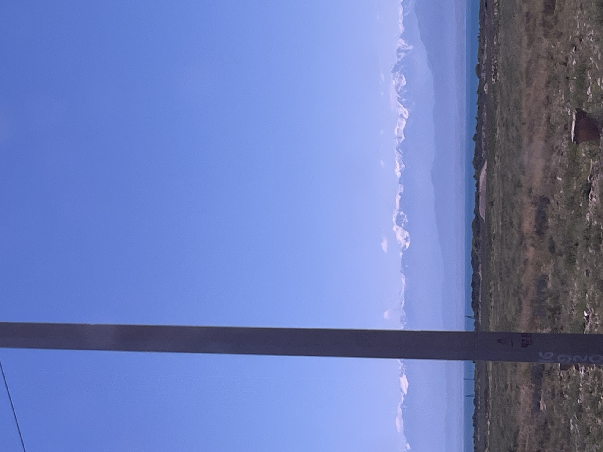 A vertical utility pole, gray and marked with some writing, stands in the foreground, slightly left of center. Beyond the pole, a vast, arid landscape stretches out, covered in dry, green and brown grasses and low shrubs. A body of water, likely a lake, is visible in the middle ground, reflecting the bright blue sky. In the far distance, a range of majestic mountains with snow-capped peaks dominates the horizon, their outlines softened by atmospheric haze. The sky above is a clear, vibrant blue, devoid of clouds.