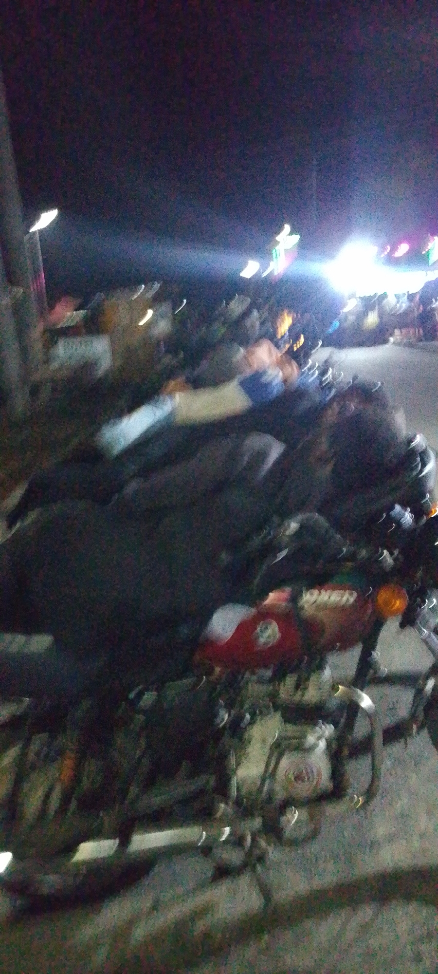The image, taken at night in what appears to be Lokoja, Nigeria, shows several motorcycles parked along a street. The motorcycle closest to the viewer is a red "Boxer" model. Streetlights and other illuminated signs suggest a commercial or public area. There are blurry indications of people standing in the background. The overall visibility is low.