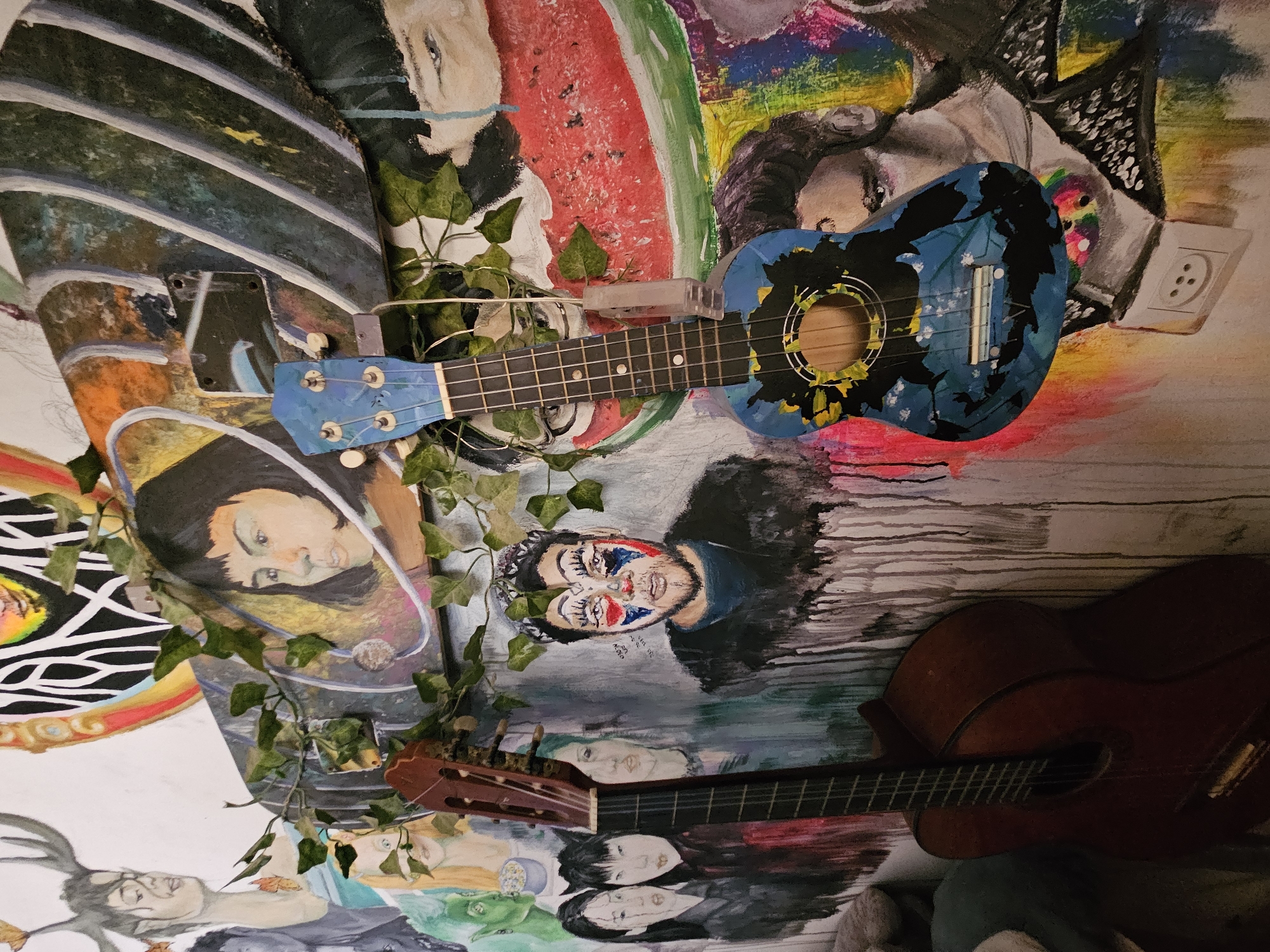 The image depicts an indoor scene, likely a wall decorated with an eclectic mix of paintings and graffiti. Two musical instruments, a guitar and a ukulele, are prominently featured, leaning against the wall. The guitar, a classical acoustic with a dark wood finish, is positioned on the left. To its right, a blue ukulele, adorned with a painted yellow and black floral or abstract design, hangs from the wall. The wall itself is a canvas for various artworks, including portraits of people with striking facial expressions and makeup, a depiction of a watermelon slice, and abstract splashes of color and dripping paint. Green ivy vines are draped across the instruments and the wall art, adding a touch of organic texture to the scene. The overall impression is one of artistic expression and a lived-in, bohemian atmosphere.
This scene is located in Rishon LeZion, Israel, specifically at the "Ziv Street" music school.