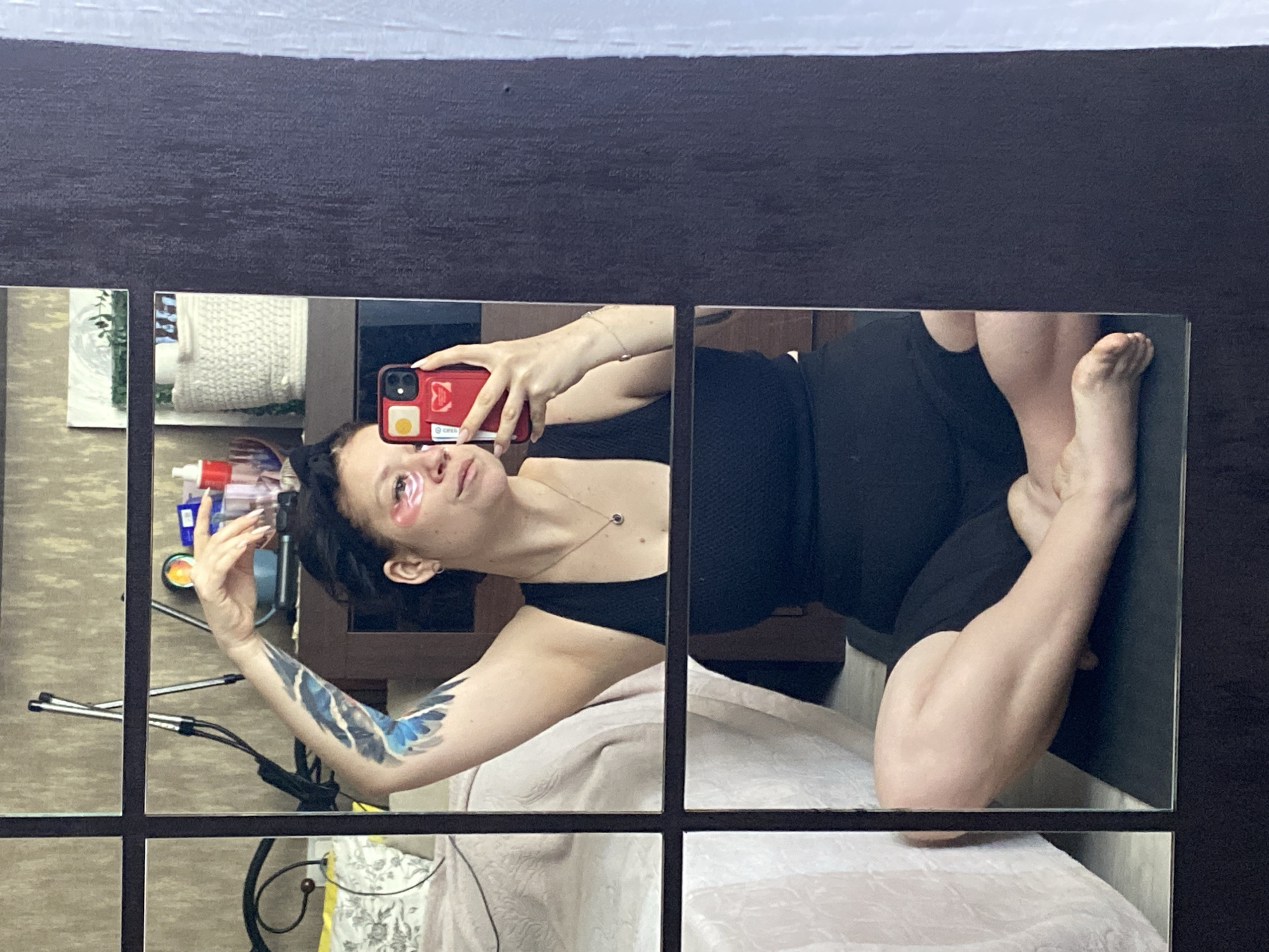 A woman is taking a selfie in a grid-patterned mirror. She is lying on a bed with a light-colored blanket. She is wearing a black tank top and dark pants. She has eye patches under her eyes and a blue bird tattoo on her upper arm. She is holding a red phone. There is a shelf with various items on it in the background. The location is unknown.