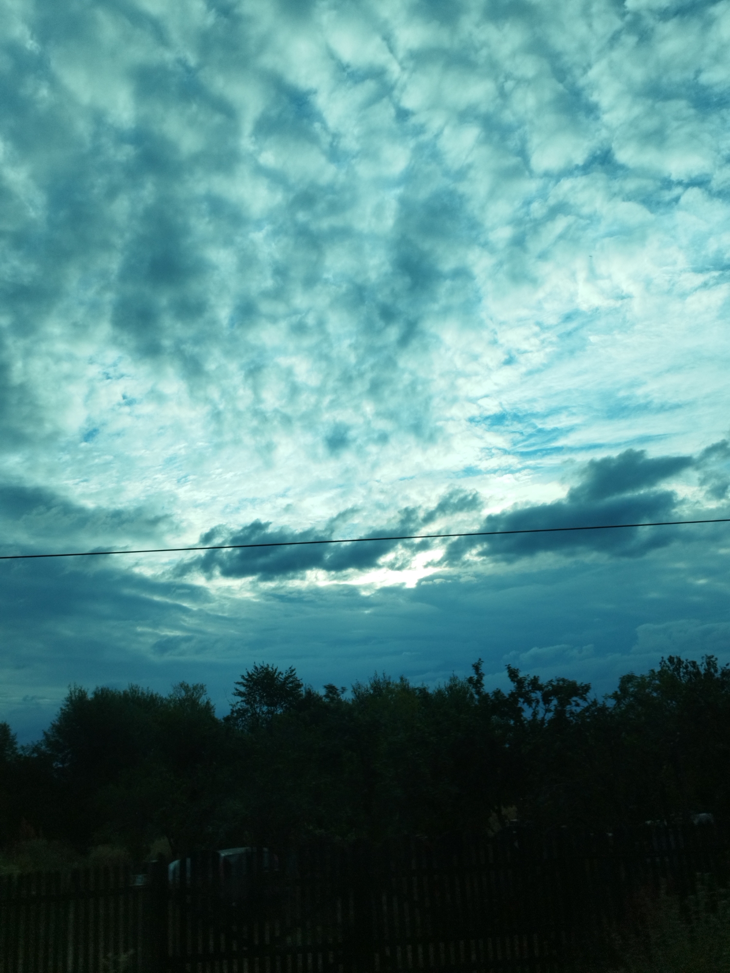 The image depicts a cloudy sky with a turquoise hue above a silhouette of trees and a fence. A dark horizontal line, likely a power line, traverses the mid-section of the sky. The lower portion of the image is significantly darker, showing the outlines of dense foliage and a wooden fence. In the background, obscured by darkness, there are hints of structures or vehicles behind the fence. The scene appears to be an outdoor view, possibly from a residential area, in Cuza Vodă, Romania, given the context provided. The lighting suggests either early morning or late evening, with the sun partially visible through breaks in the clouds.