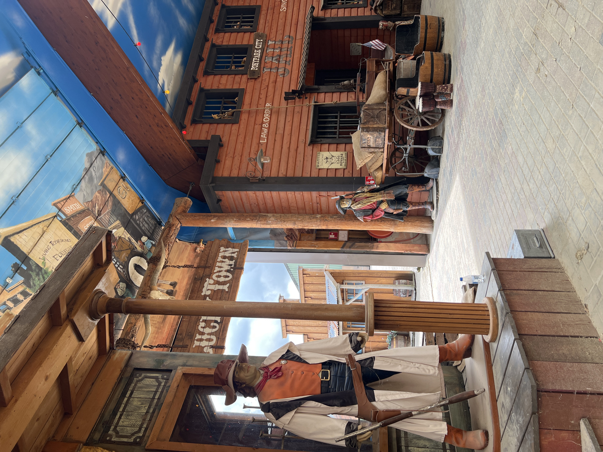 The image depicts an indoor Western-themed attraction, likely in the city of Collendoorn, Netherlands. In the foreground, a mannequin dressed as a cowboy stands on a wooden platform, holding a rifle. The mannequin wears a brown hat, an orange vest, a long cream-colored coat, and brown boots, with a gun belt and holster. Behind the cowboy mannequin, a sign reads "LUCKY TOWN".
To the right, a wooden building facade mimics a Wild West jail. It is painted red and features barred windows. A sign above the entrance reads "POSTAGE CITY JAIL". A wagon laden with sacks and crates is parked in front of the jail building, alongside several wooden barrels and two drums. Another mannequin dressed as a cowboy stands near the wagon, holding a rifle.
The ceiling is decorated to resemble a blue sky with clouds and painted buildings, giving the impression of an outdoor scene within an enclosed space. The ground is paved with cobblestones. The overall scene is designed to evoke a historical Western town setting.