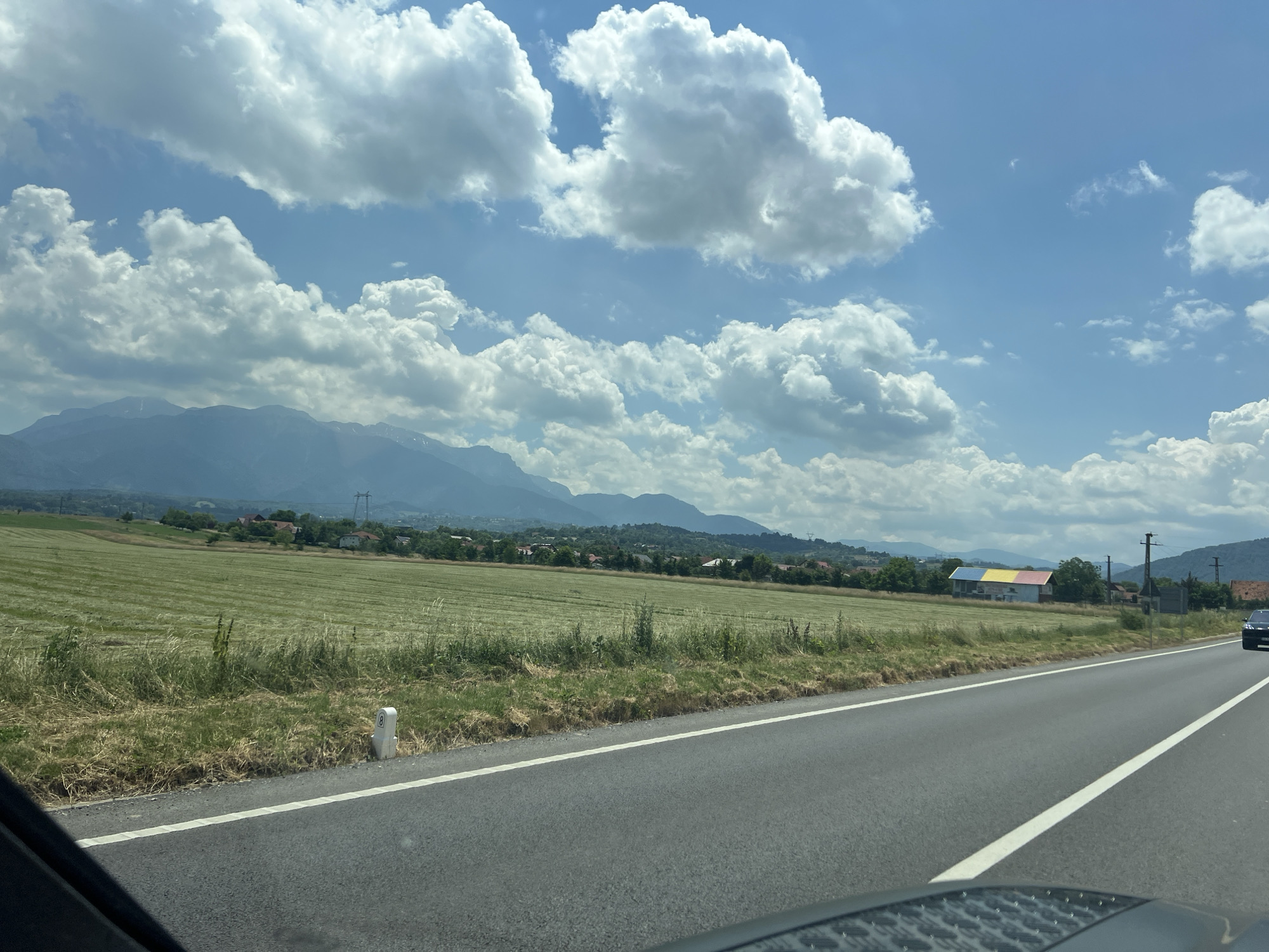 A wide shot captures a scenic view of a rural landscape under a bright, partly cloudy sky. In the foreground, a gray asphalt road with white lane markings runs diagonally across the frame from the bottom right corner. The road appears to be a two-lane highway with a single white line separating the lanes. To the left of the road, a dry, grassy verge with a white marker post stands out. Beyond the verge, a vast, green field stretches towards a distant line of trees and houses. The field has a textured pattern, suggesting it has been recently mowed. In the mid-ground, a cluster of buildings is visible, one of which is distinctively painted with horizontal stripes of blue, yellow, and red. Further in the distance, a mountain range forms a hazy silhouette against the sky. The sky is a vibrant blue, dotted with large, fluffy white cumulus clouds. The overall impression is one of a serene, sunlit day in the countryside.