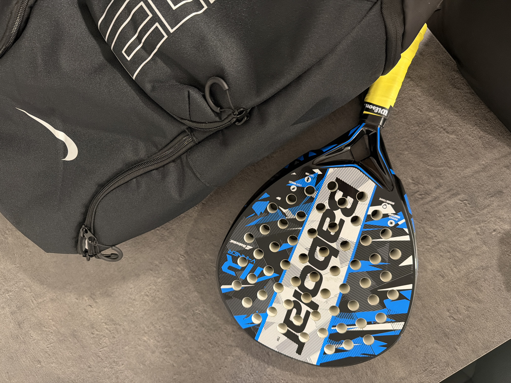 An overhead, eye-level shot shows a black Nike backpack and a blue and black Babolat padel racket. The backpack is positioned to the right of the frame, partially obscuring the racket. The racket lies horizontally across the bottom of the frame, its yellow grip extending towards the bottom right corner. The background is a textured gray surface, possibly a stone floor or fabric. The scene appears to be indoors, as there is no indication of outdoor elements. The lighting is even, suggesting artificial illumination. There are no people or animals visible in the image.