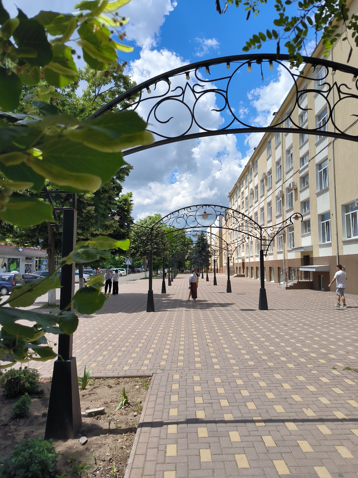 In Tiraspol, Moldova, this image captures a sunlit pedestrian walkway lined with decorative arched structures. The walkway is paved with light brown and yellow rectangular bricks laid in a herringbone pattern, creating a visually appealing texture. Flanking the walkway are areas of dirt with sparse green vegetation, interspersed with black metal posts supporting the arches. Several black metal arches, adorned with intricate scrollwork and small light bulbs, extend along the pathway, casting subtle shadows.
On the left side of the walkway, a multi-story building with a beige facade and numerous white-framed windows dominates the scene. Air conditioning units are visible mounted below many of the windows. A few trees with lush green foliage are planted along this side of the walkway, adding to the urban greenery.
On the right side of the walkway, the building continues, with a wider, slightly recessed entrance featuring a grey awning and handrails leading to stairs. Further down, a person in a white t-shirt and grey shorts is walking away from the camera.
In the middle ground of the walkway, three individuals are observed. Two people in business attire, a man and a woman, are standing and conversing. To their right, a woman in a white top and a reddish-brown skirt is walking away from the camera. In the distance, more people are scattered along the walkway, and cars are parked on the adjacent street.
The sky above is a vibrant blue, dotted with fluffy white clouds, indicating a pleasant day. In the foreground, large green leaves from a tree obscure a portion of the view, framing the scene with organic elements.