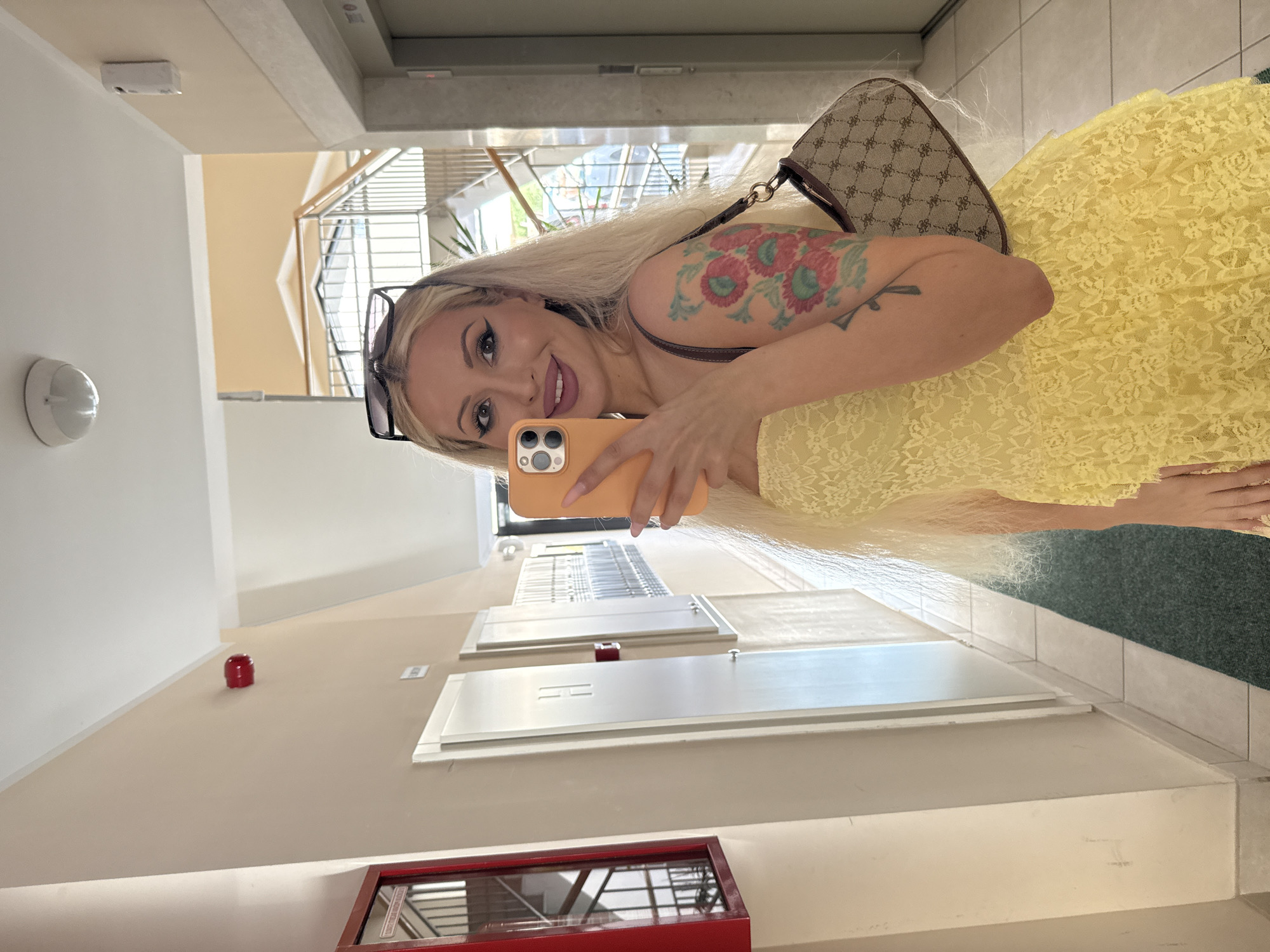 A blonde woman is captured in a selfie, taken with an orange smartphone held in her right hand. She wears sunglasses atop her head, features winged eyeliner and lined lips, and exhibits a colorful floral tattoo on her left upper arm, with a smaller dark tattoo below it. Her attire consists of a yellow, tiered lace dress, and she carries a small, brown, patterned shoulder bag on her left shoulder. The scene is an indoor hallway with beige walls and a white ceiling, featuring a dark green mat on the floor transitioning to lighter tiles. On the left wall, a red fire alarm strobe light is visible above a red-framed glass cabinet that reflects a staircase. Multiple white utility panels are mounted on the wall, one prominently featuring an embossed "H". To the right, an elevator door is partially visible. In the background, beyond an archway, a multi-story staircase with metal railings can be seen. A white circular ceiling light fixture and a white wall-mounted air freshener unit are also present.