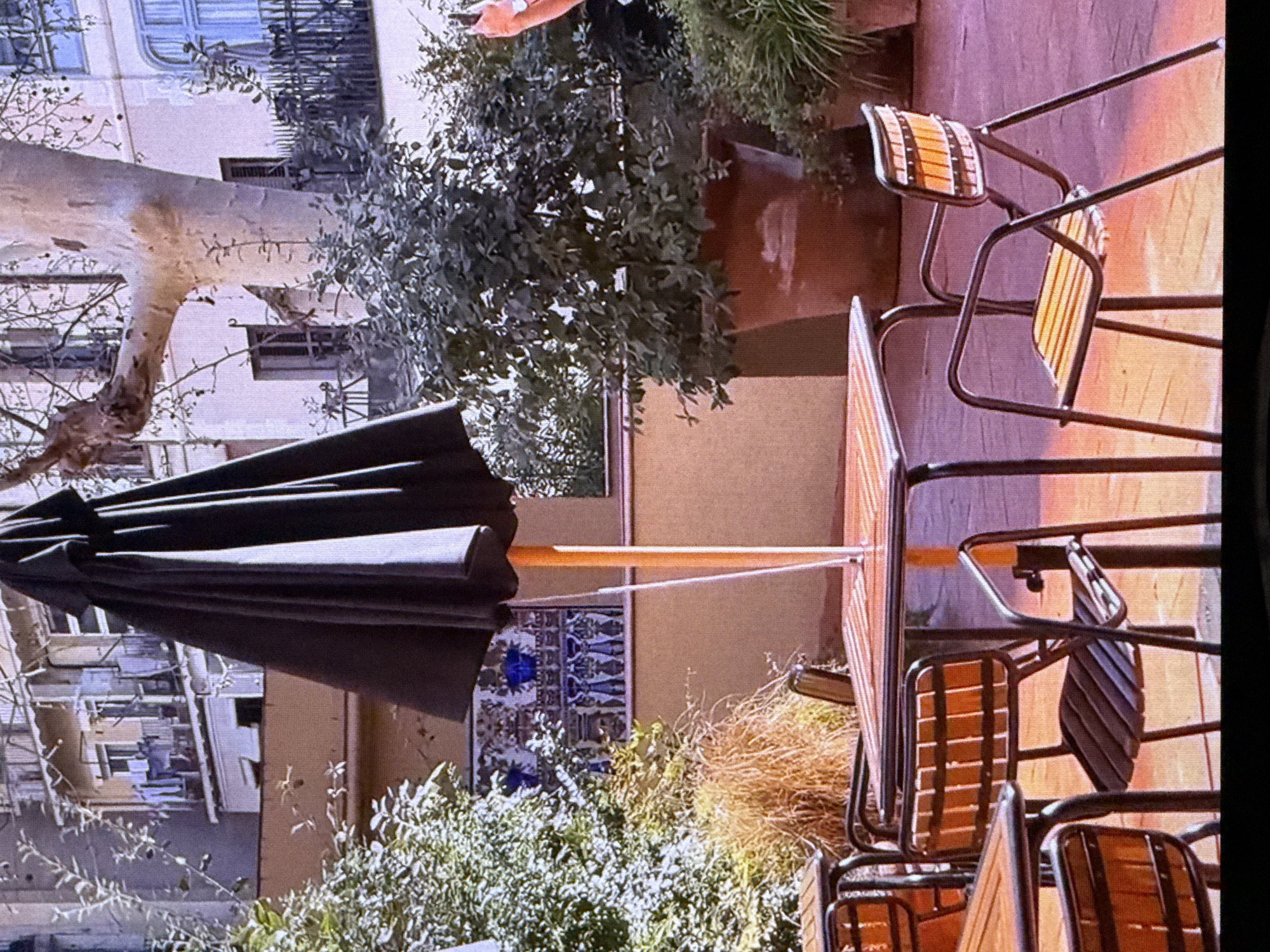 An outdoor cafe scene is depicted with tables and chairs set up. A large black umbrella is open, providing shade over a wooden table. Several chairs with black metal frames and wooden slatted seats and backs are arranged around the tables. In the background, there are buildings with balconies and windows, some of which have laundry hanging out. Lush green foliage and a large potted plant are visible, adding to the ambiance of the outdoor space. The ground is paved with a reddish-orange material. The overall impression is that of a serene and inviting setting for dining or relaxing outdoors. This scene is not located in Dubai, United Arab Emirates.