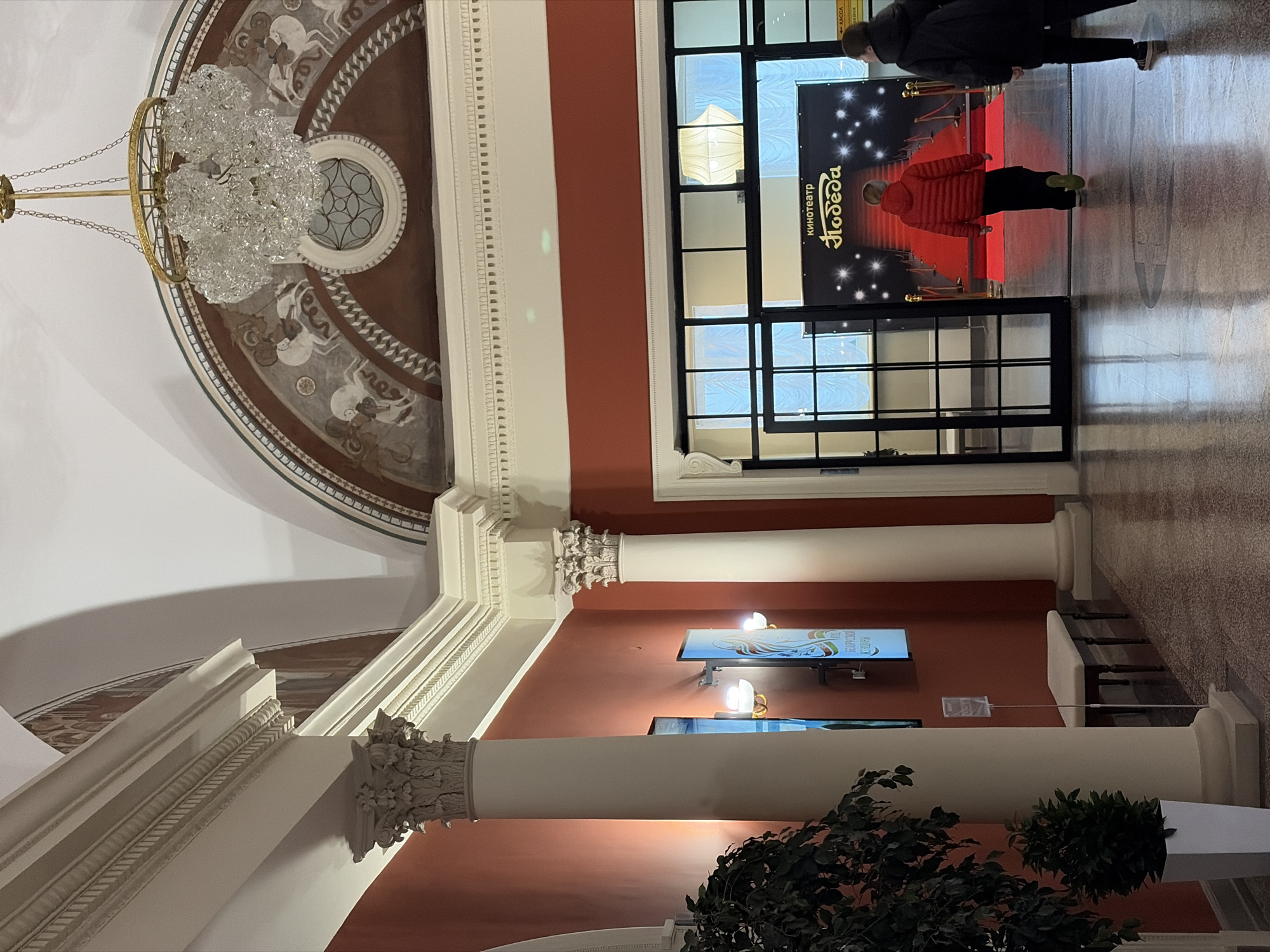 The image shows the interior of the Pobeda Cinema in Volgograd, Russia. A large crystal chandelier hangs from a patterned ceiling. A red carpet leads into the cinema. Two people are visible: one wearing a dark coat and the other in a red jacket. The pillars supporting the building feature ornate capitals. Two television screens hang on the wall between the columns. A potted plant is placed on the floor.