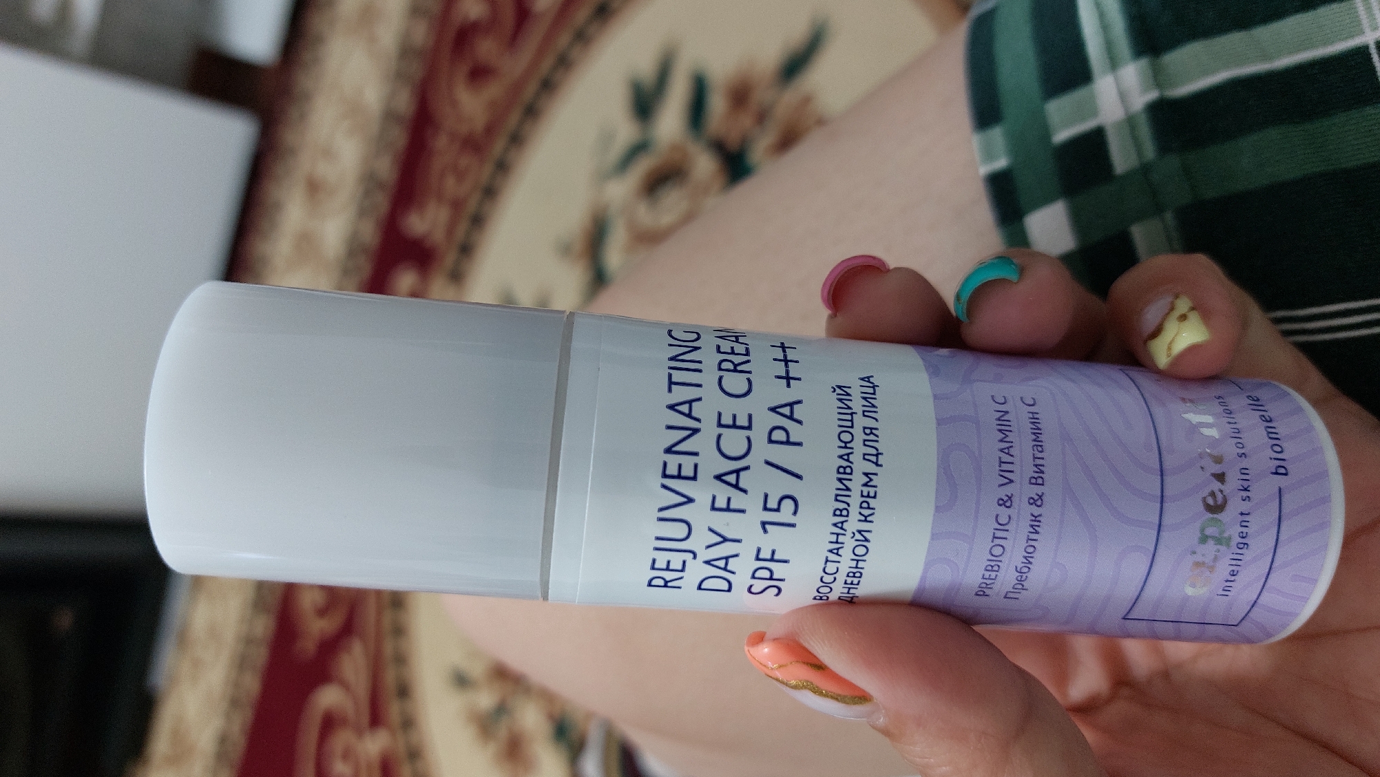 A person is holding a white and lavender bottle of "REJUVENATING DAY FACE CREAM SPF 15 / PA +++" with Russian text below. The bottle has a white pump top and is partially obscured by the person's hand, which has painted nails. The background appears to be a patterned carpet or rug. No specific location in Kara-Balta, Kyrgyzstan is identifiable from the image.