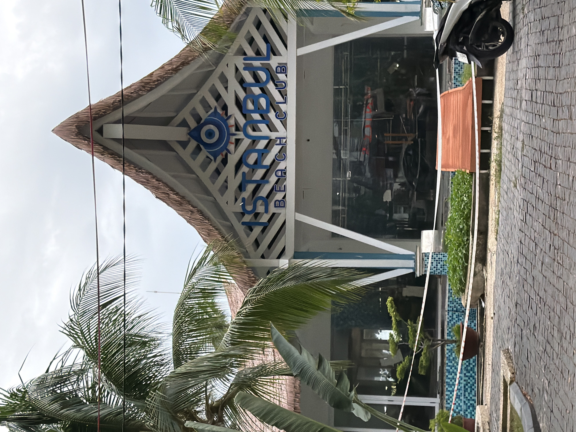 The image shows the entrance of the "Istanbul Beach Club" in Nha Trang, Vietnam. The building features a triangular thatched roof and a white facade with blue trim. The words "ISTANBUL BEACH CLUB" are prominently displayed in blue lettering above large glass windows. Palm trees frame the building, and a paved walkway leads up to the entrance. In the foreground, there is a motorcycle parked on the walkway, and a reddish-orange bench is visible in front of the building. The area appears to be an outdoor establishment with a swimming pool or decorative water feature visible behind red and white caution tape. The sky is overcast.