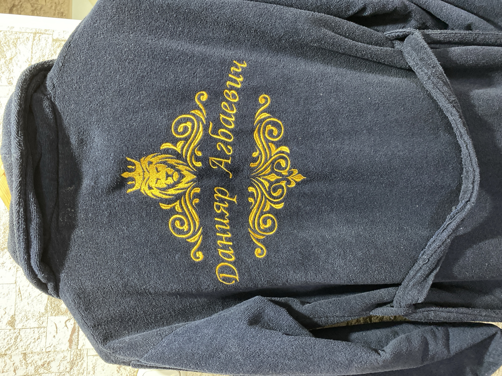 The image shows the back of a dark blue terrycloth robe. Embroidered on the back of the robe in gold thread is a crest featuring a lion's head wearing a crown, surrounded by decorative scrolls. Below the crest, the name "Данияр Агбаевич" is embroidered in elegant script. The robe is tied with a sash at the waist. The background appears to be a textured wall, possibly a brick or stone pattern, with a light colored wall to the right. The scene is indoors. There are no people or animals visible in the image. The robe is likely a personal item, possibly a gift or a custom-made piece. The embroidery suggests a personal touch, perhaps for a special occasion or as a sign of luxury. The location is specified as Бескөл ауылдық округі, Kazakhstan.