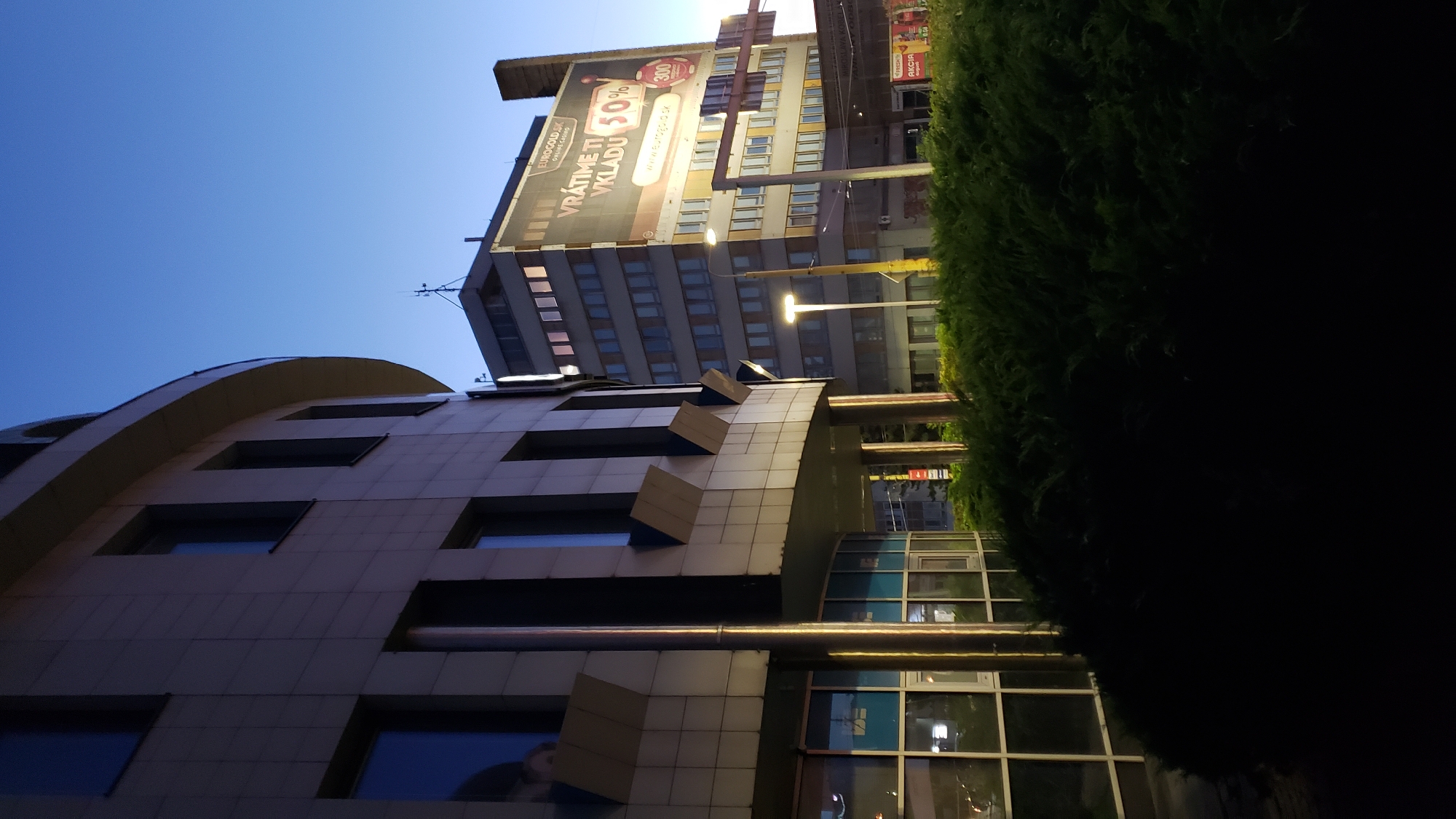 The image depicts an outdoor scene at dusk in Prešov, Slovakia, with two modern office buildings and a dense hedge in the foreground. The building on the left is curved and features a light-colored facade with a grid of rectangular windows. The building on the right is taller and rectangular, covered in a large advertisement for "EUROGOLD.SK ONLINE CASINO." This advertisement prominently displays "VRÁTIME TI VKLADU 50%" (We'll return your deposit 50%) and a website address, "www.eurogold.sk." Streetlights illuminate the area, and the sky is a deep blue, indicating twilight. The dense, dark green hedge obscures the lower portion of the scene and the ground. The overall impression is one of an urban environment at the end of the day.