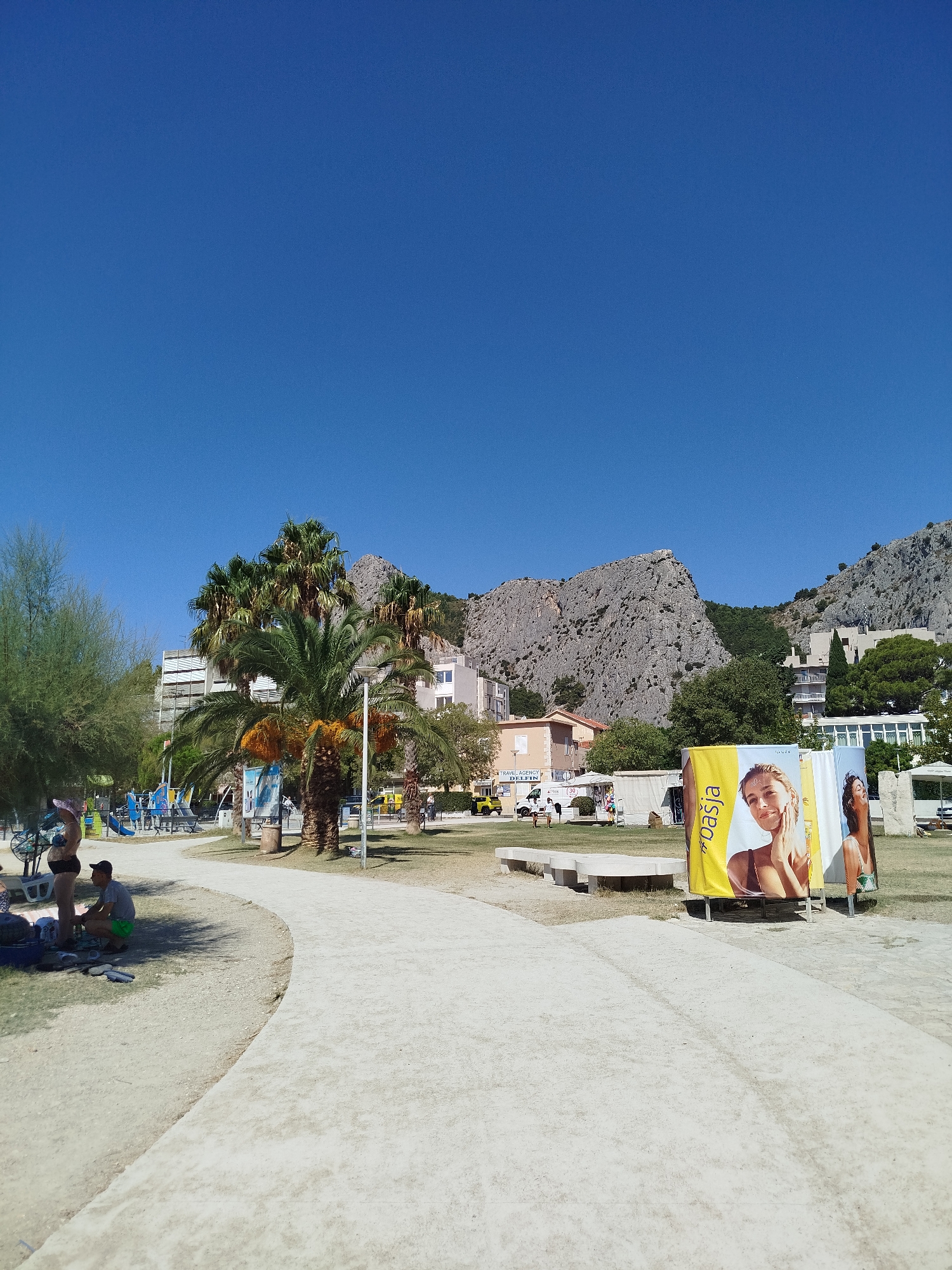The image depicts a sunny outdoor scene in Omiš, Croatia, likely a beach or park area near the coast. The prominent feature is a wide, light-colored paved path curving through the foreground. To the left of the path, a sandy area is visible with several people. A woman in a bikini and a hat stands near a bicycle, while a man in shorts crouches nearby. There are bags and flip-flops on the sand.
Further into the scene, a playground with blue and yellow equipment is set up. Several palm trees, some bearing orange fruit, are scattered throughout the landscape, with lush green foliage. Behind the trees and playground, buildings, some appearing to be hotels or apartments, are visible. A notable geological feature, a rugged, rocky mountain or cliff face, looms in the background, characteristic of the Omiš region.
On the right side of the path, large vertical banners are displayed. One banner features a woman's face with the text "pasja" written vertically in yellow and white. Another banner shows another woman. The ground is mostly covered in grass and sand. In the distance, a yellow vehicle is parked, and other people can be seen milling about. The sky is a clear, bright blue. The overall impression is a pleasant, active day in a tourist-oriented location.