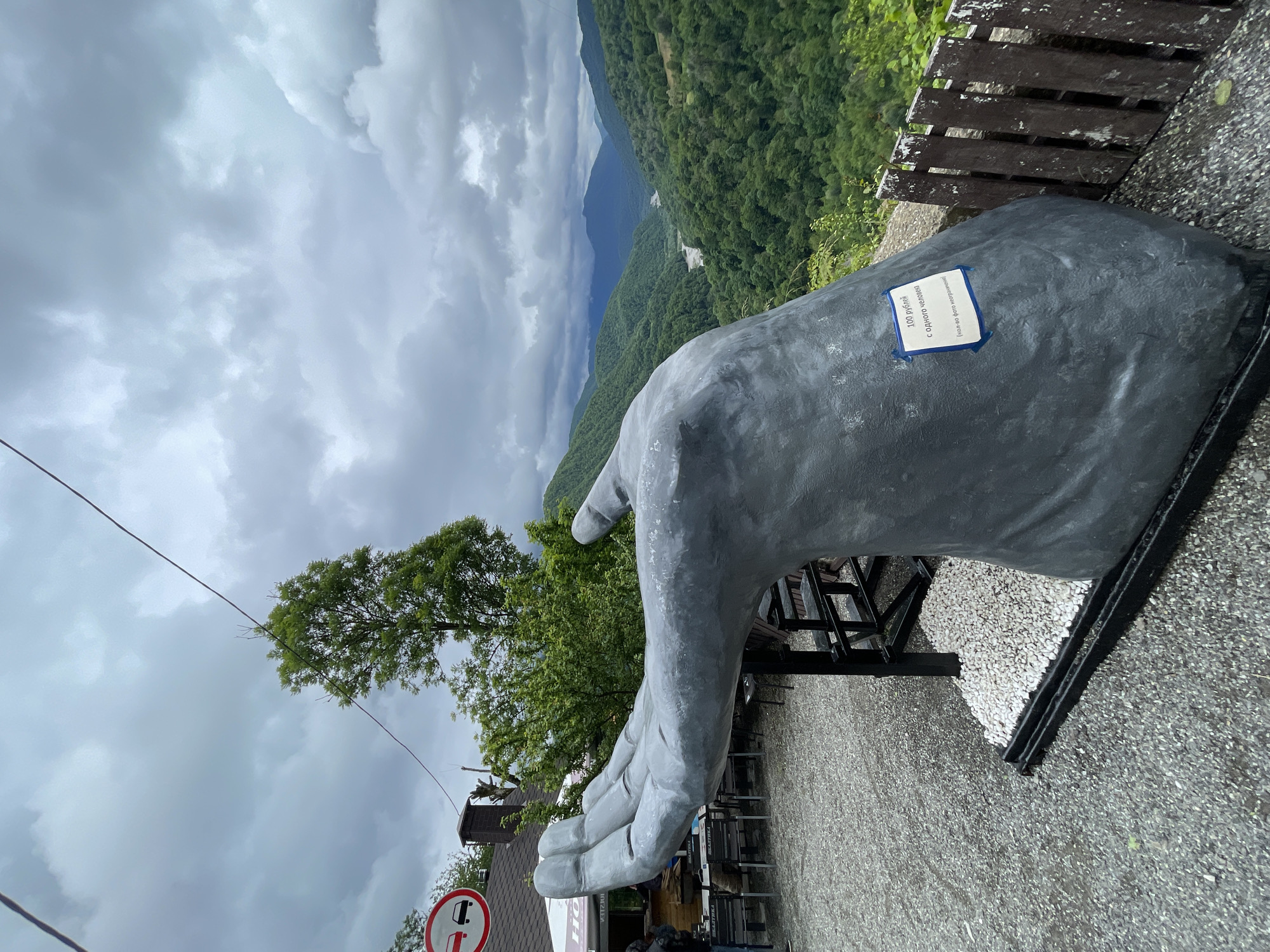 The image shows a large, gray, sculpture of a hand near what appears to be a scenic overlook, possibly in Khartsyz Pervyi, Russia. The hand has a blue sign affixed to it. In the background is a landscape of rolling, green hills and mountains under an overcast sky. A wooden fence and a gravel surface are visible near the sculpture. A tree and a building can be seen near the base of the sculpture, and a traffic sign is located at the bottom-left of the image. There are tables and chairs in the area as well.