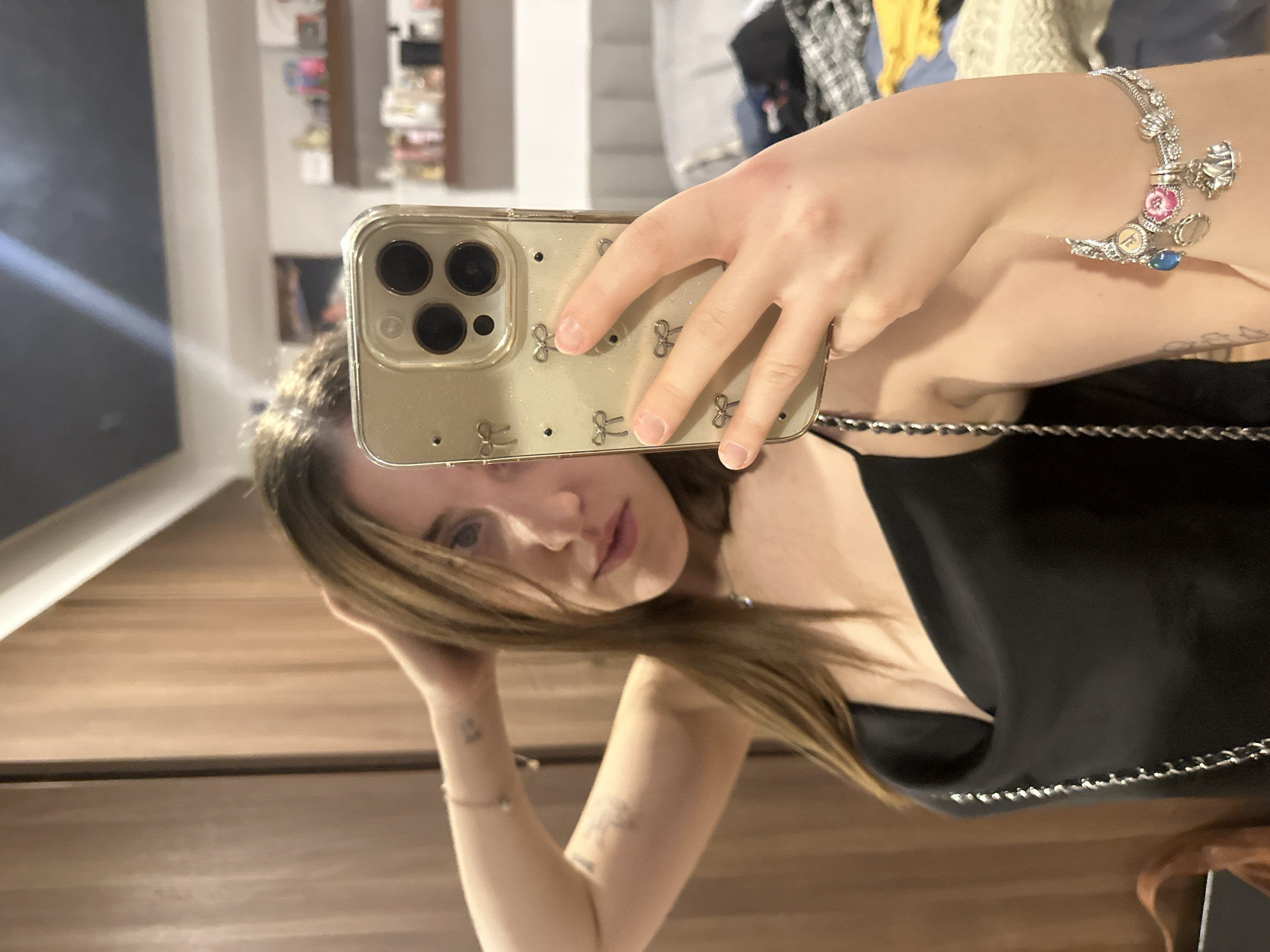 A woman is taking a selfie in a mirror. She is holding a smartphone with a clear case decorated with bows in her right hand. She has a bracelet on her right wrist. She is wearing a black top with a chain strap. She has light brown hair. The background shows a wall with a wooden cabinet and a shelf with various items. The location is possibly in Huixquilucan de Degollado, Mexico, based on the prompt request.