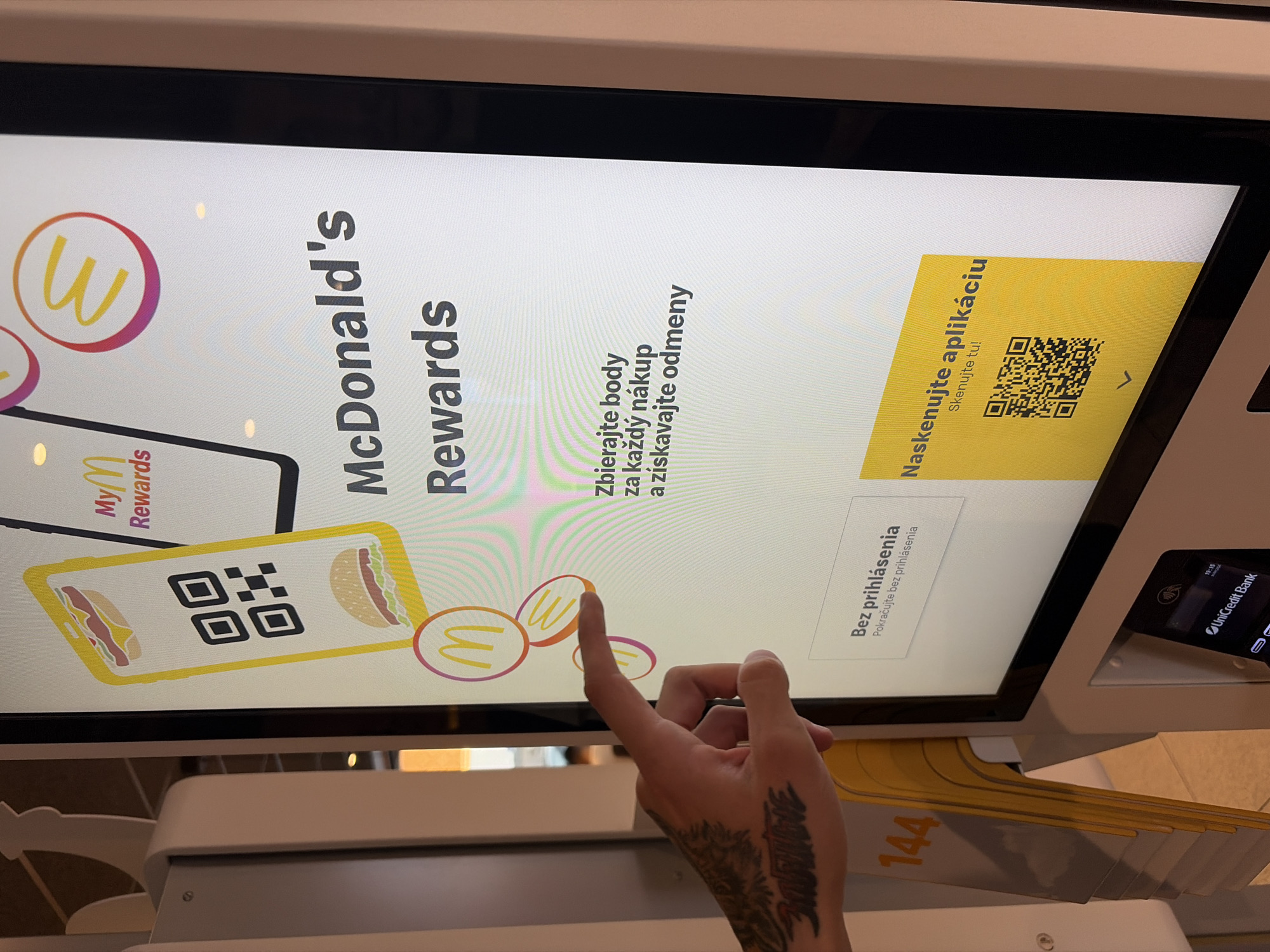 An individual's tattooed hand is visible in the foreground, with their index finger extended towards a touchscreen display at a McDonald's restaurant in Nitra, Slovakia. The screen displays information about McDonald's Rewards, including a graphic of a smartphone with a QR code and the text "Zbierajte body za každý nákup a získavajte odmeny" (Collect points for every purchase and get rewards). Below this, there is an option to "Bez prihlásenia" (Without logging in) and "Naskenujte aplikáciu" (Scan the application) with a QR code. The background shows the interior of the restaurant, including a stack of yellow order trays and part of a self-order kiosk.