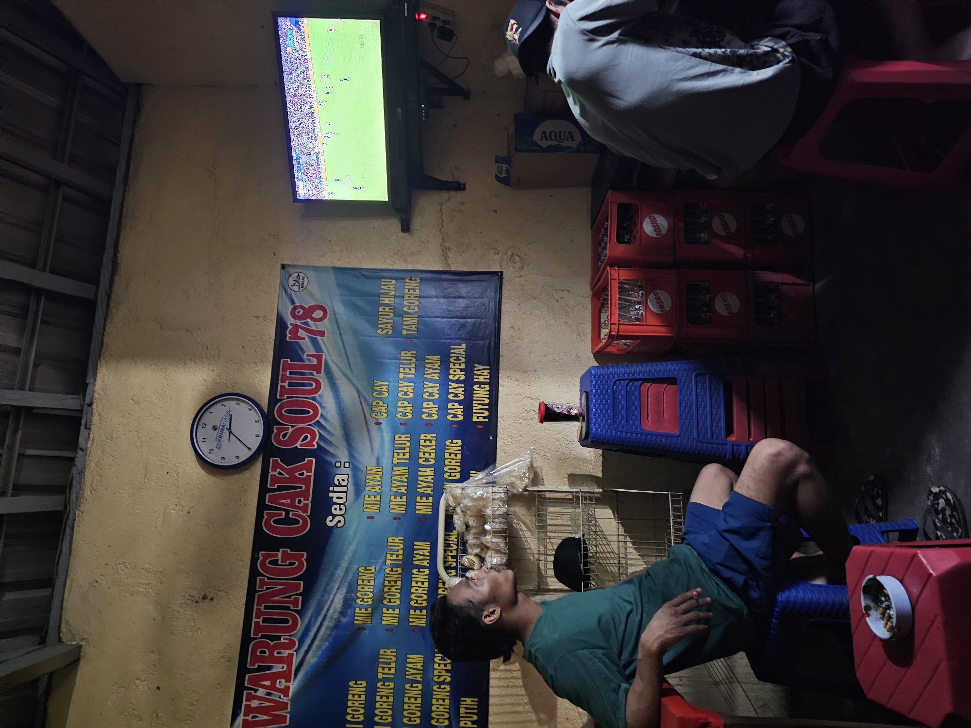 The image shows an informal open-air eatery in Denpasar, Indonesia. In the background, a TV screen displays a football match. A large banner for "Warung Cak Soul 78" is mounted on a yellow wall, listing menu items like "Mie Goreng," "Mie Ayam," and "Cap Cay." A clock on the wall reads approximately 5:25.
Two men are present. One is sitting on a red stool, his back to the camera. The other is sitting on a blue plastic chair, wearing a green t-shirt and blue shorts. He is smoking a cigarette. A red ashtray sits next to him.
Several crates of "Sosro" brand beverages are stacked near the men, alongside a box of "Aqua" water bottles. Cooking equipment can be seen behind the seated man, including a metal rack with packages of snacks or ingredients.