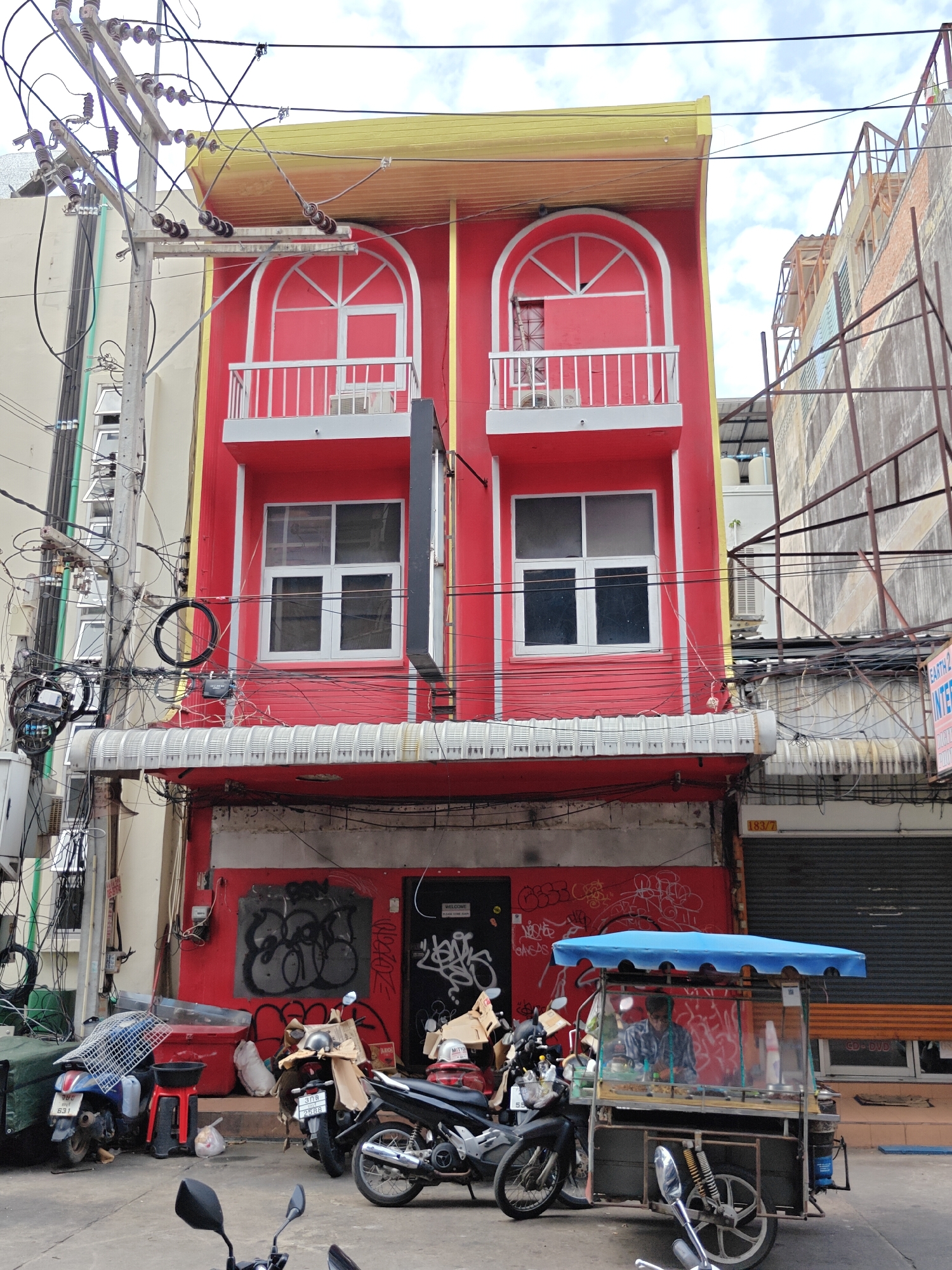 This is a daytime, eye-level, exterior, full shot of a vibrant red building on a city street in Pattaya City, Thailand. The building has a yellow roof and is adorned with white trim around its arched windows and balconies. The ground floor features graffiti on the red wall and a black door. In front of the building, a street vendor operates a food cart with a blue canopy, serving a customer. Several motorcycles are parked nearby, along with a red plastic stool and various bags and containers. The street is cluttered with electrical wires and poles, indicative of an urban environment. The background shows other buildings and a cloudy sky.