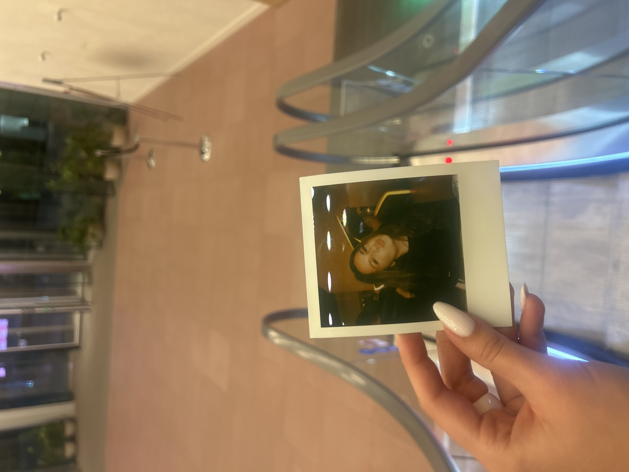 A hand with long, almond-shaped white fingernails holds a square Polaroid photograph in front of a modern building interior. The photo shows a woman with long brown hair looking at the camera. The building interior features a curved metal and glass structure, a pale pink tiled wall, and a window with plants and lights visible outside. The building could potentially be located in the city of London, United Kingdom.