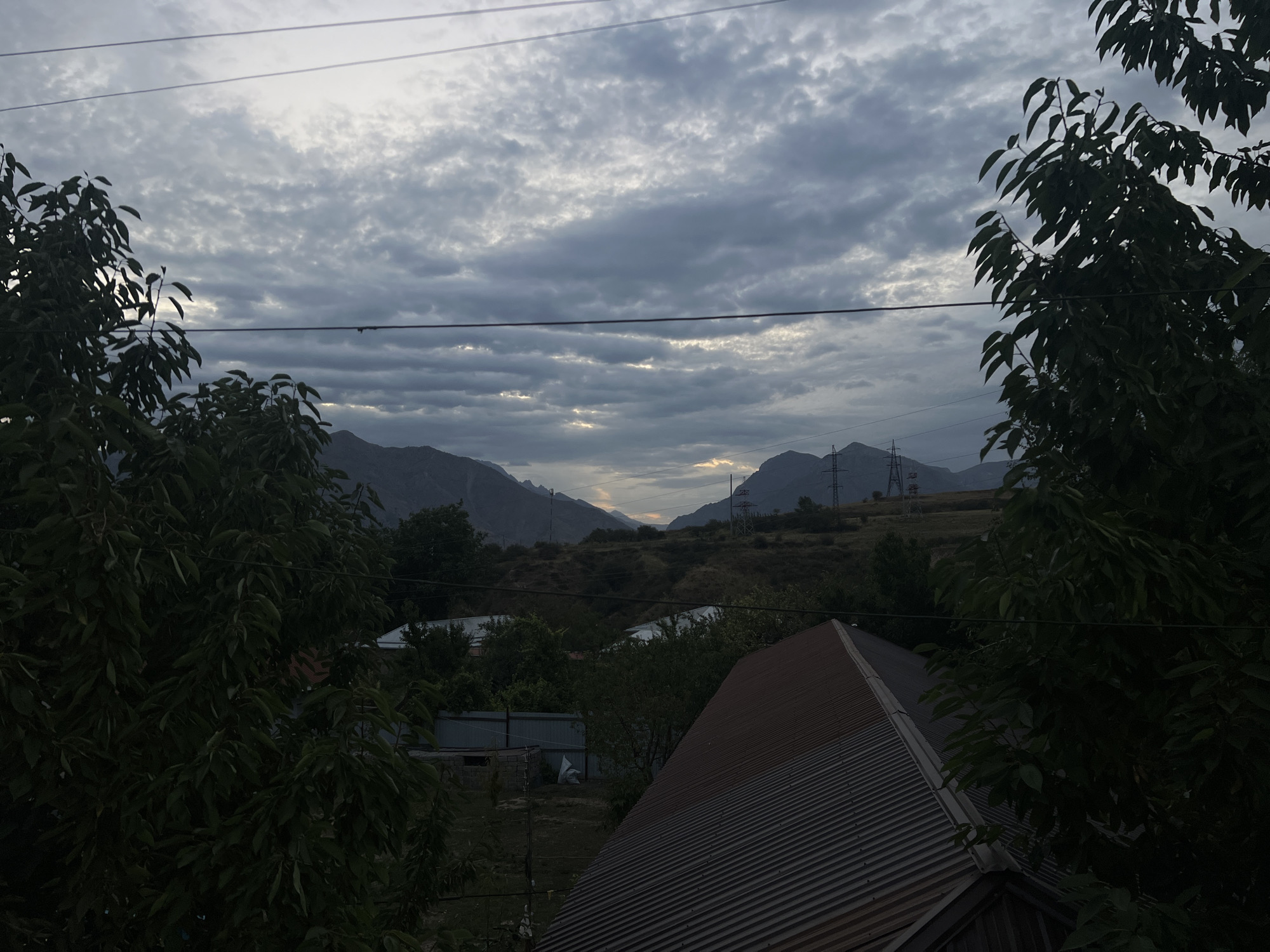 This is a horizontal, medium-shot, eye-level, exterior photograph taken in Yusuphana, Uzbekistan, of a cloudy, gray sky over a mountainous landscape. In the foreground, on the left and right, are dark green leafy trees. In the middle ground, there are several buildings with metal roofs. In the background, there are mountains. Two power lines are visible crossing the sky.