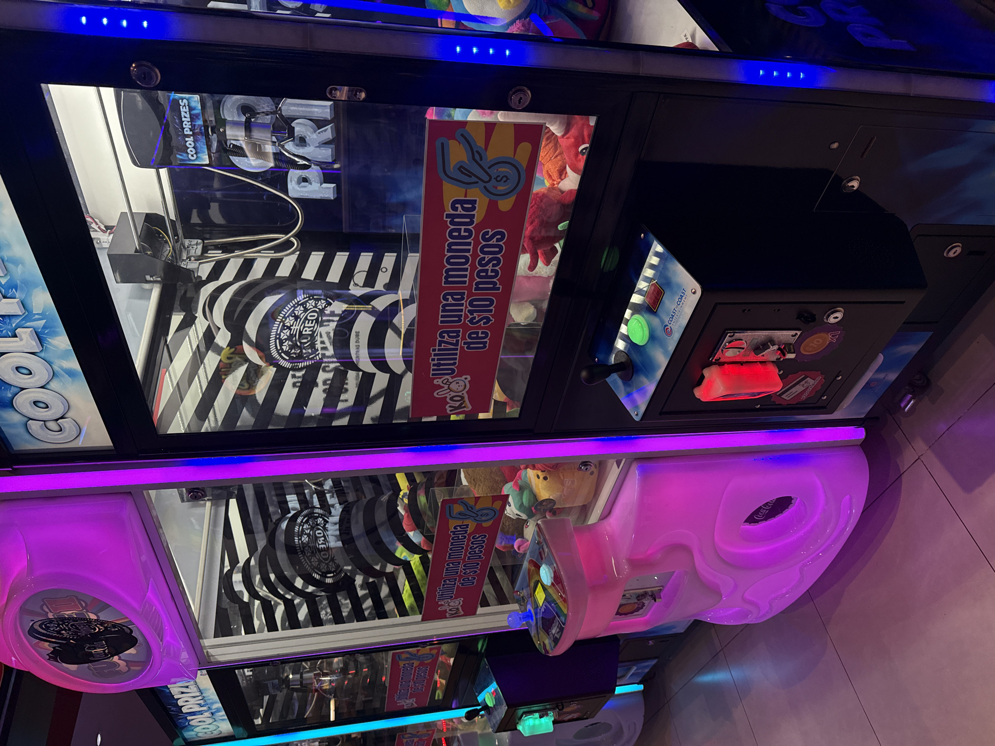 This indoor shot shows two "Cool Prizes" claw machines. The top machine is decorated with blue lights, while the bottom one is illuminated with pink. The machines have prizes inside, including what appear to be Oreo-branded merchandise and plush toys. A sign in Spanish on both machines reads "Utiliza una moneda de $10 pesos," indicating the cost to play is 10 pesos. A "Coast-to-Coast" logo is visible near the controls of the top machine. A "Coca-Cola" logo is also visible near the lower claw machine. Given the price in pesos and the inclusion of a Coast-to-Coast logo (a company with presence in Mexico), it can be speculated that this scene is located in the city of Puebla, Mexico.