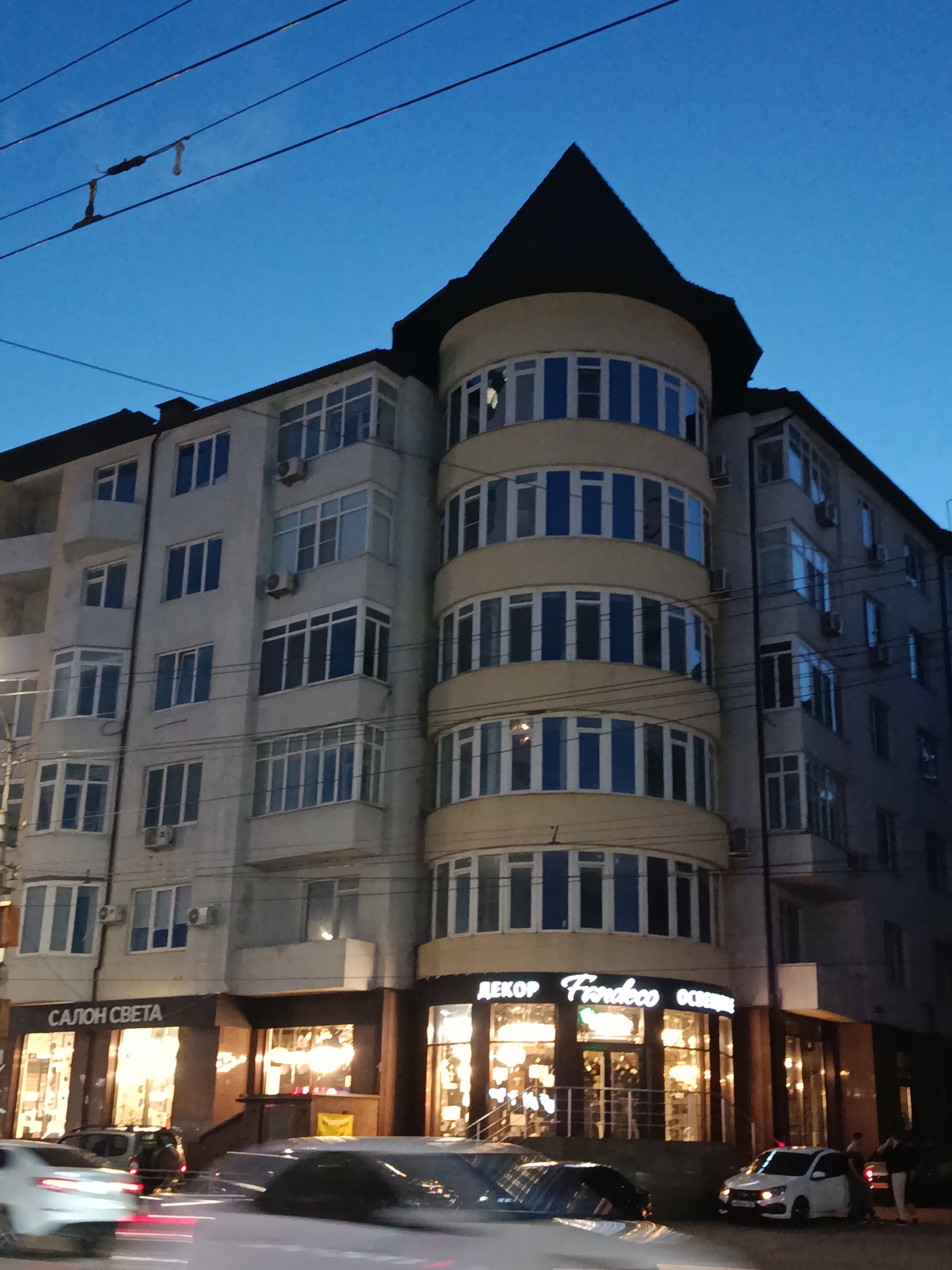 The image shows a building in Saratov, Russia, at dusk or early evening. The building is multi-story with a rounded section featuring a conical roof. The facade has numerous windows and balconies. On the ground floor, there are commercial establishments with illuminated signs. One sign reads "САЛОН СВЕТА" (Salon of Light) in Cyrillic. Another sign, also in Cyrillic and Latin script, appears to say "ДЕКОР Frodeco ОСВЕЩЕНИЕ" (Decor Frodeco Lighting). Cars are present on the street in front of the building, some of which are in motion, indicated by motion blur. The sky is a deep blue, suggesting it is not yet fully dark. Overhead, there are power lines and possibly overhead train or tram wires.