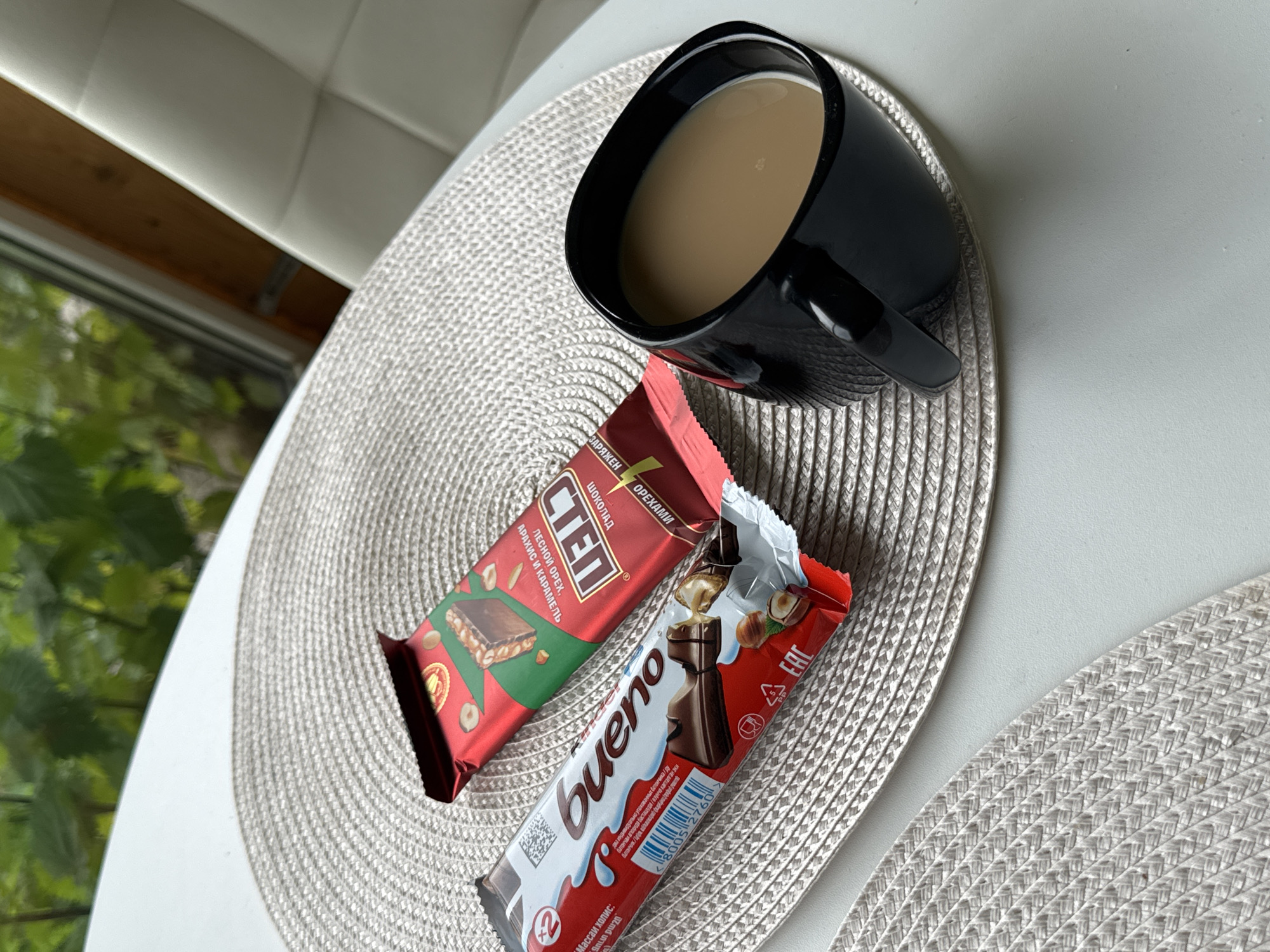 A top-down shot captures a breakfast scene with two chocolate bars and a mug of coffee. The setting appears to be indoors, possibly near a window given the glimpses of greenery outside. The light suggests it's daytime.
Dominating the frame are two chocolate bars placed on a woven placemat. The bar on the left is red and features Russian text that includes "СТЕП" and "ШОКОЛАД С ОРЕХАМИ" (Chocolate with Nuts). It also mentions "ЛЕСНОЙ ОРЕХ, АРАХИС И КАРАМЕЛЬ" (Hazelnut, Peanut, and Caramel), with an image of a chocolate bar with nuts and caramel.
To the right, and slightly open, is a Kinder Bueno bar. Its distinctive white and red packaging is visible, with the "bueno" logo prominently displayed. The bar itself shows its wafer and chocolate construction, along with whole hazelnuts.
A black mug, filled with a light brown beverage, likely coffee with milk, is positioned above the chocolates. The mug has a sleek, modern design with a thick handle. The placemat under the items is a circular, textured, light-colored weave. The overall arrangement on a plain white surface suggests a simple, everyday moment of enjoying a sweet treat with coffee. The presence of "EAC" on the Kinder Bueno packaging indicates compliance with Eurasian Customs Union standards.