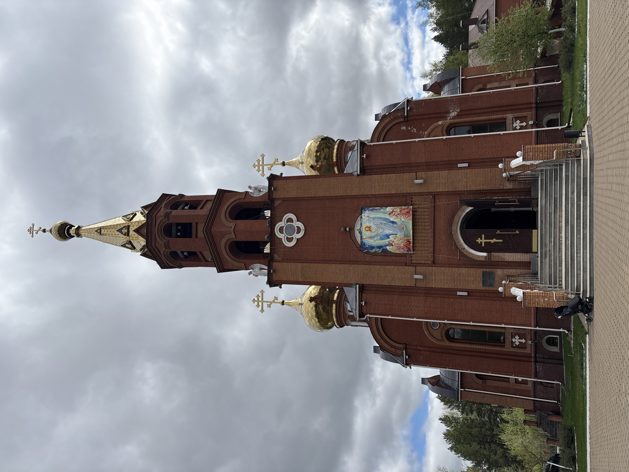 The image depicts a brick church located in the city of Chaikovsky, Russia. The church features a tall, golden-topped spire and several golden domes, each topped with a cross. A mosaic depicting a religious figure is visible on the facade. The entrance to the church is accessible via a set of stairs. The sky is overcast with heavy clouds. A stroller is parked near the entrance.