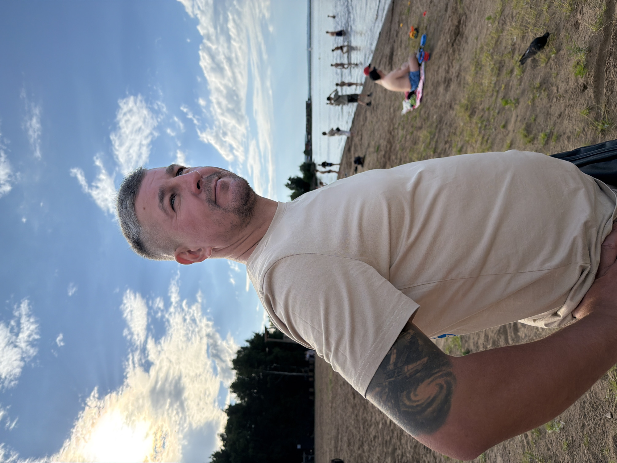 This media file depicts a beach scene with several people. The main subject is a man in the foreground, with his head and shoulder visible. He is looking upwards, and his expression is neutral. He has short, grey hair and a beard. He is wearing a light-colored t-shirt and has a tattoo on his arm.
In the background, there are other people on the beach and in the water. Some are standing near the water's edge, while others are further back on the sand. There are also some children's toys scattered on the sand. A bird is visible on the sand in the mid-ground.
The scene appears to be set during the day, with a blue sky and some clouds. The sun is shining, as indicated by the bright light and some lens flare. The overall mood is relaxed and casual, suggesting a day at the beach. The presence of trees in the background and the body of water indicate a natural outdoor setting. The crops provided show a close-up of the man's face and sky, a wider view of the beach with people, a view of the sand with toys and a bird, and a close-up of the man's arm and t-shirt.