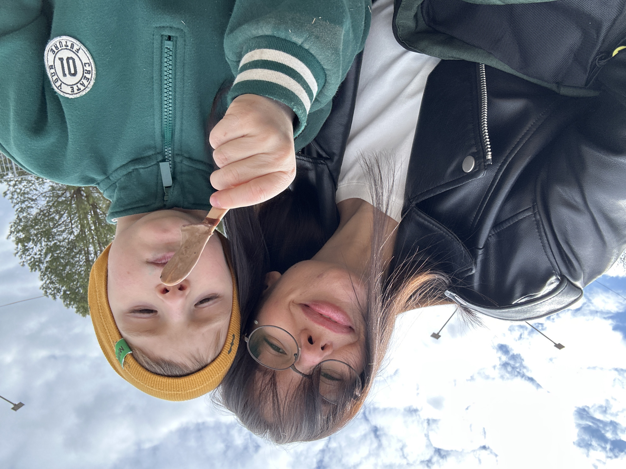 The image is an upside-down selfie of a woman and a child holding an ice cream treat. The woman is wearing glasses, a white shirt, and a black leather jacket. The child has a yellow beanie and a green jacket with white stripes on the sleeve and a "10 Future" patch on the chest. The background shows a cloudy sky and a blurred view of trees. Based on these details, it is impossible to determine the specific location in the city of Chaikovsky, Russia.