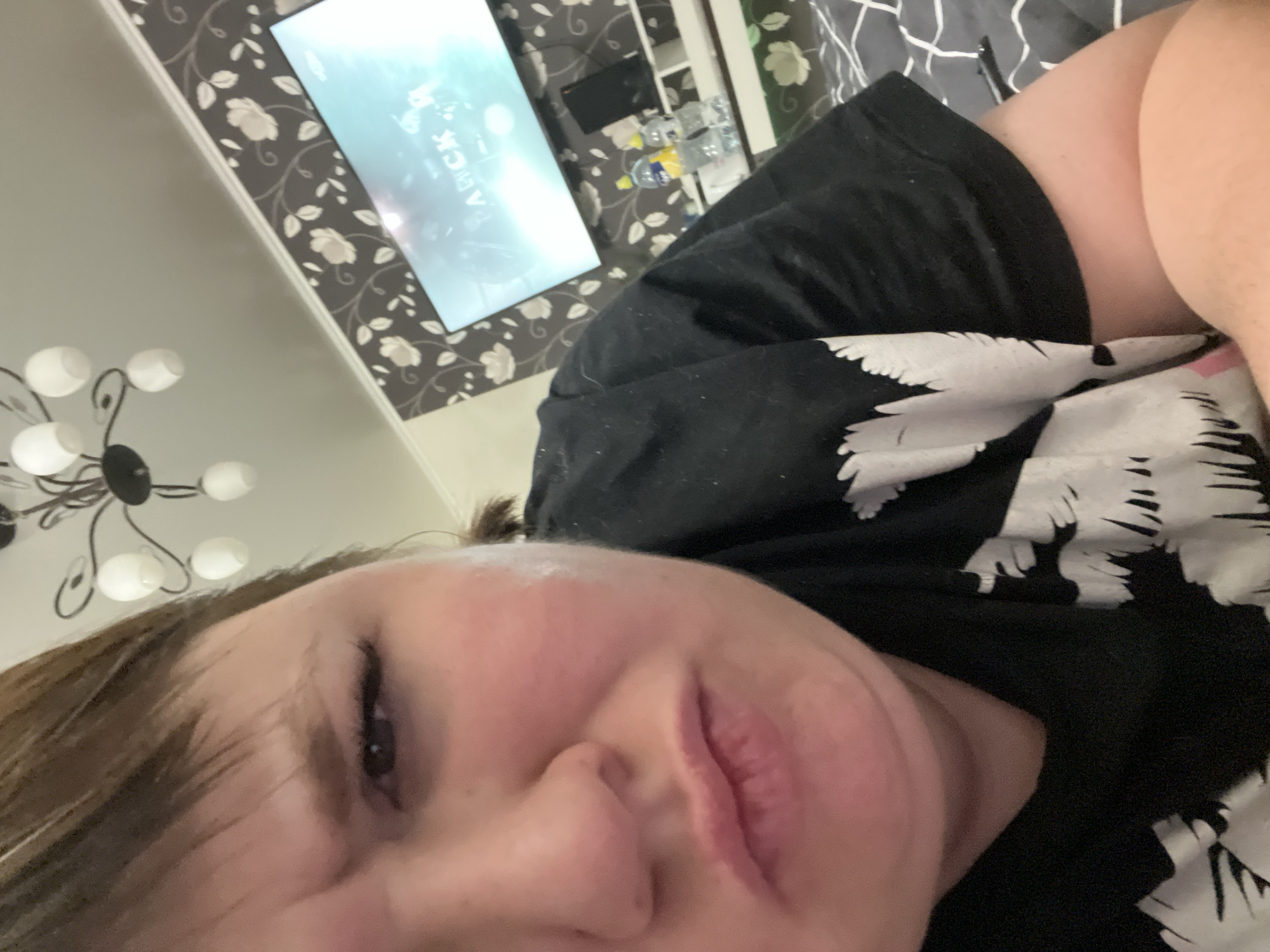 A selfie-style photograph shows a woman lying down, facing the camera. She has fair skin, brown hair, and is wearing a black shirt with a white pattern. A television screen is visible in the background, displaying the word "AРCK". The background also includes patterned wallpaper, a ceiling light fixture, and a table with multiple water bottles. No information in the image suggests the location is the city of Chaikovsky, Russia.