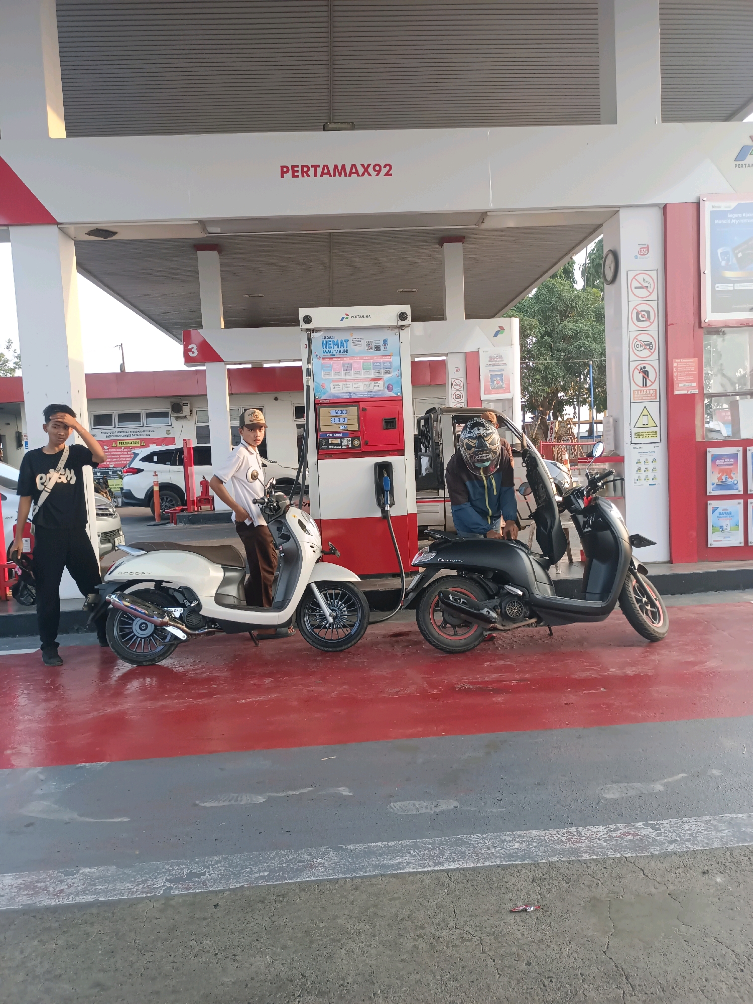 The image depicts a gas station in Subang, Indonesia, with three individuals and two motorcycles. Two young men are standing near a Pertamax 92 gas pump. One, wearing a black t-shirt and black pants, stands to the left, with his right hand raised to his forehead. A white scooter is parked in front of him. To the right of the pump, another young man, dressed in a white polo shirt and brown pants, sits on a black scooter. He is wearing a baseball cap. A third individual, wearing a helmet and a blue and brown jacket, is leaning over the black scooter. The gas station features red and white branding, including the "PERTAMAX92" logo. Various signs and stickers are visible on the gas pump and surrounding structures, indicating prohibitions like no smoking and no photography. The ground is a combination of red and grey asphalt. In the background, a white SUV is parked, and trees are visible beyond the gas station canopy. The lighting suggests it is daytime, likely late afternoon due to the shadows.