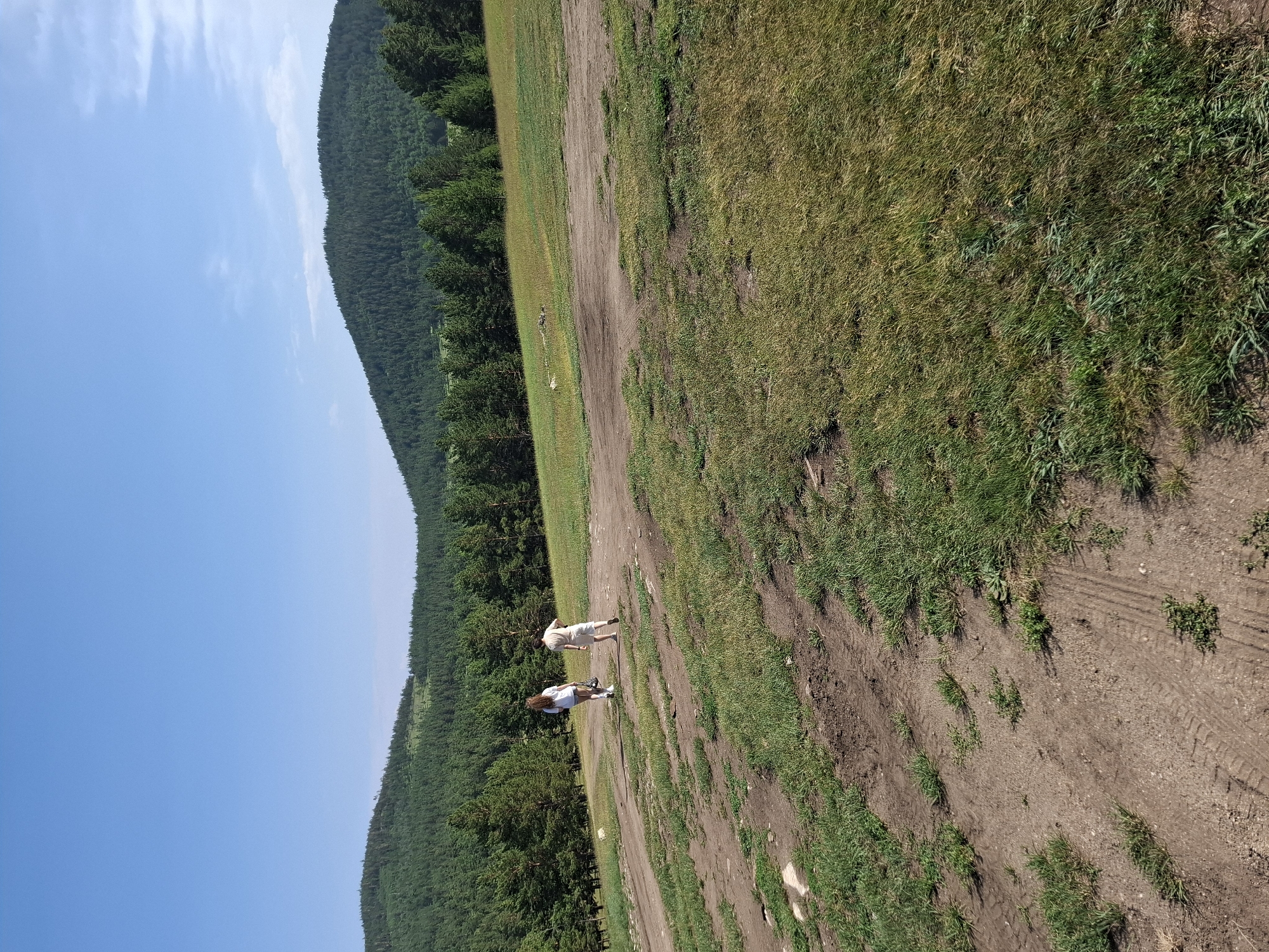A bright, sunny day in Bugul'deika, Russia, features two individuals walking away from the camera on a dirt path in a grassy, hilly landscape. The path is characterized by tire tracks and sparse green vegetation. In the background, a dense forest of pine trees covers rolling hills, leading up to a clear blue sky with a few wispy clouds. The individual on the left, wearing a white t-shirt and shorts, has long, reddish-brown hair. The person on the right is wearing a light-colored t-shirt and shorts. They appear to be walking together across the open, natural terrain.