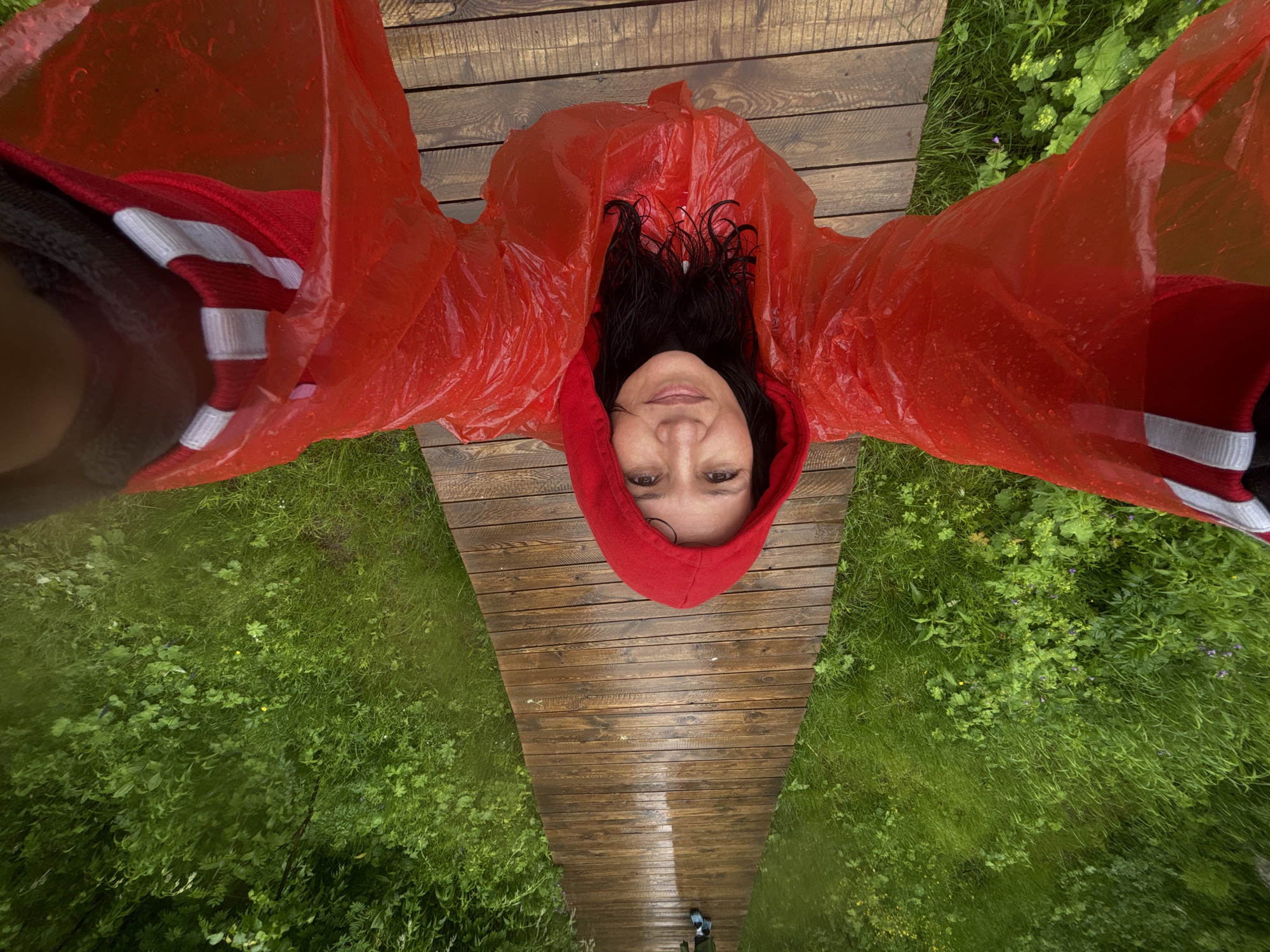 A woman wearing a red hooded raincoat stands on a wet wooden boardwalk surrounded by lush green vegetation. The boardwalk extends into the distance, where another person in green pants and blue shoes is visible. The scene appears to be an outdoor setting, possibly a park or trail, in Shumikhinskii, Russia, during a rainy day.