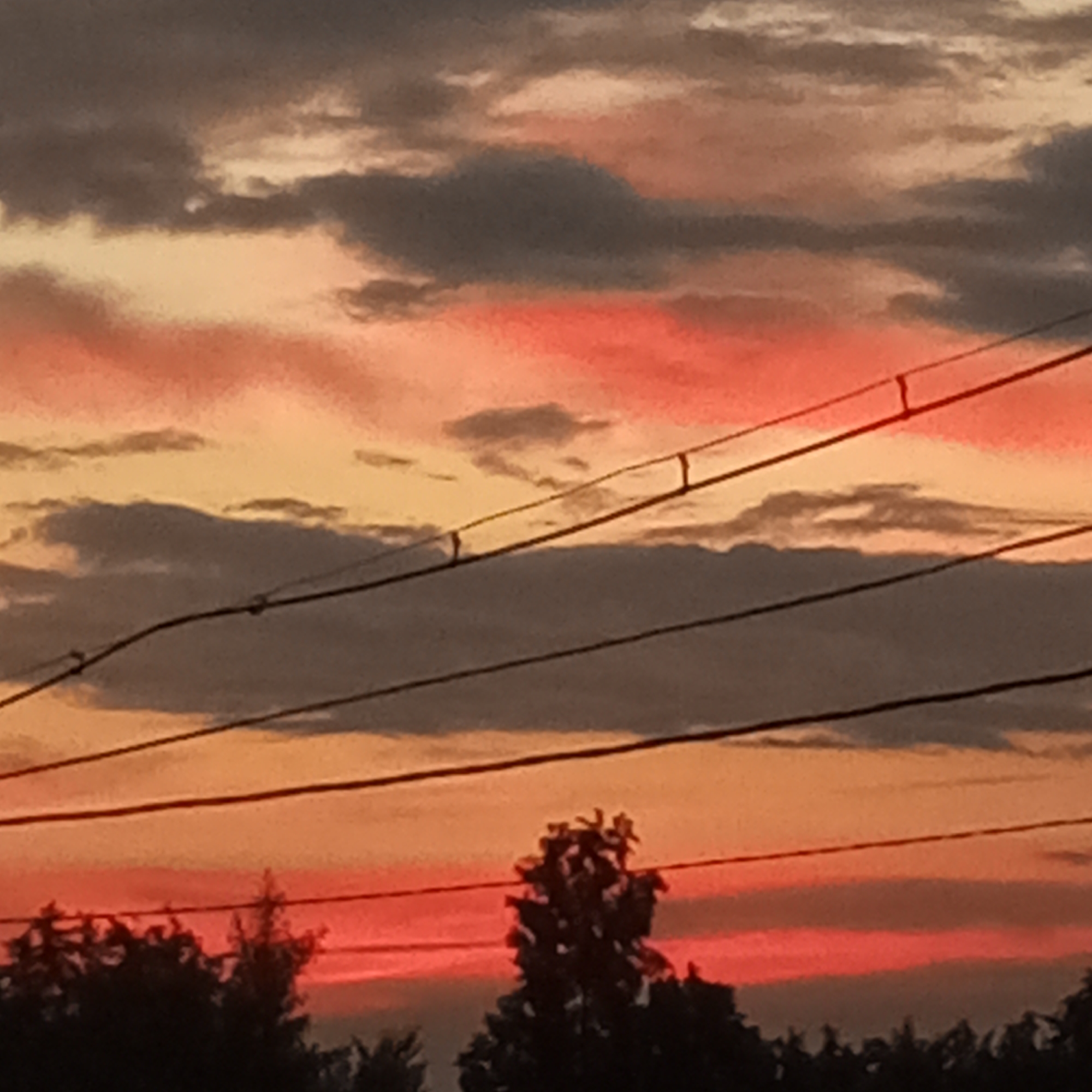 The image shows a sunset scene with a dramatic sky filled with clouds. The sky displays a gradient of colors, from dark grey and muted tones at the top to vibrant orange and red hues near the horizon. Several power lines are visible, stretching horizontally across the frame and creating a layered effect. Silhouetted against the colorful sky at the bottom are the tops of trees, suggesting a natural or suburban environment. The location in Dyurtyuli, Russia, adds a geographical context to this atmospheric view.
