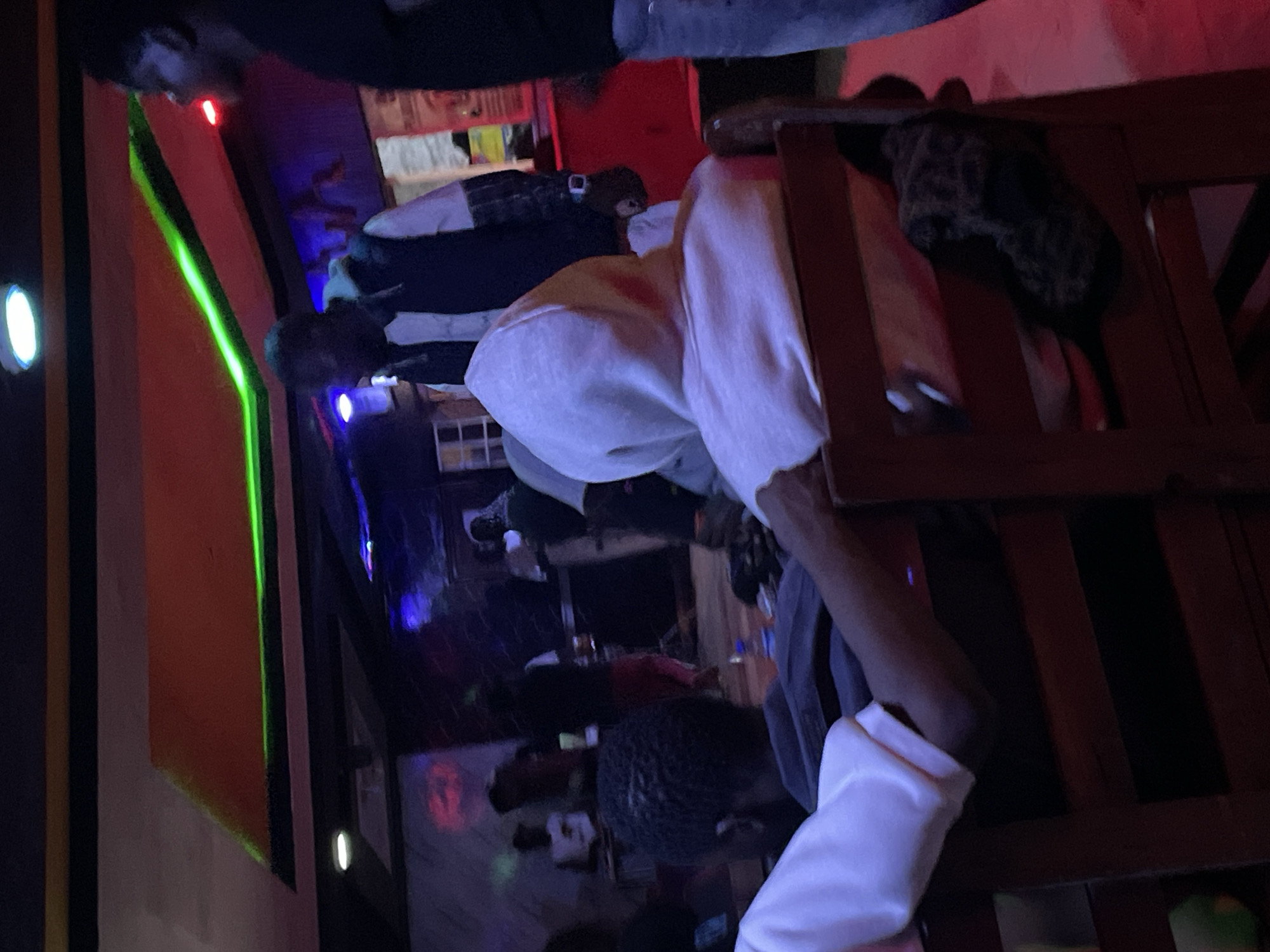 The image depicts an indoor scene, possibly a bar or lounge, in Durumi, Nigeria, characterized by dim lighting and neon accents. The environment features a suspended ceiling with an orange panel illuminated by green neon lights. Several individuals are present, some seated and others standing. In the foreground, a person wearing a white hoodie is seated, their back to the camera, with their arm extended. To their left, another individual is also seated, their head turned away. In the background, several other people are visible, including one man wearing a jacket over a plaid shirt and another individual partially obscured. The overall atmosphere suggests a social gathering in a dimly lit establishment.