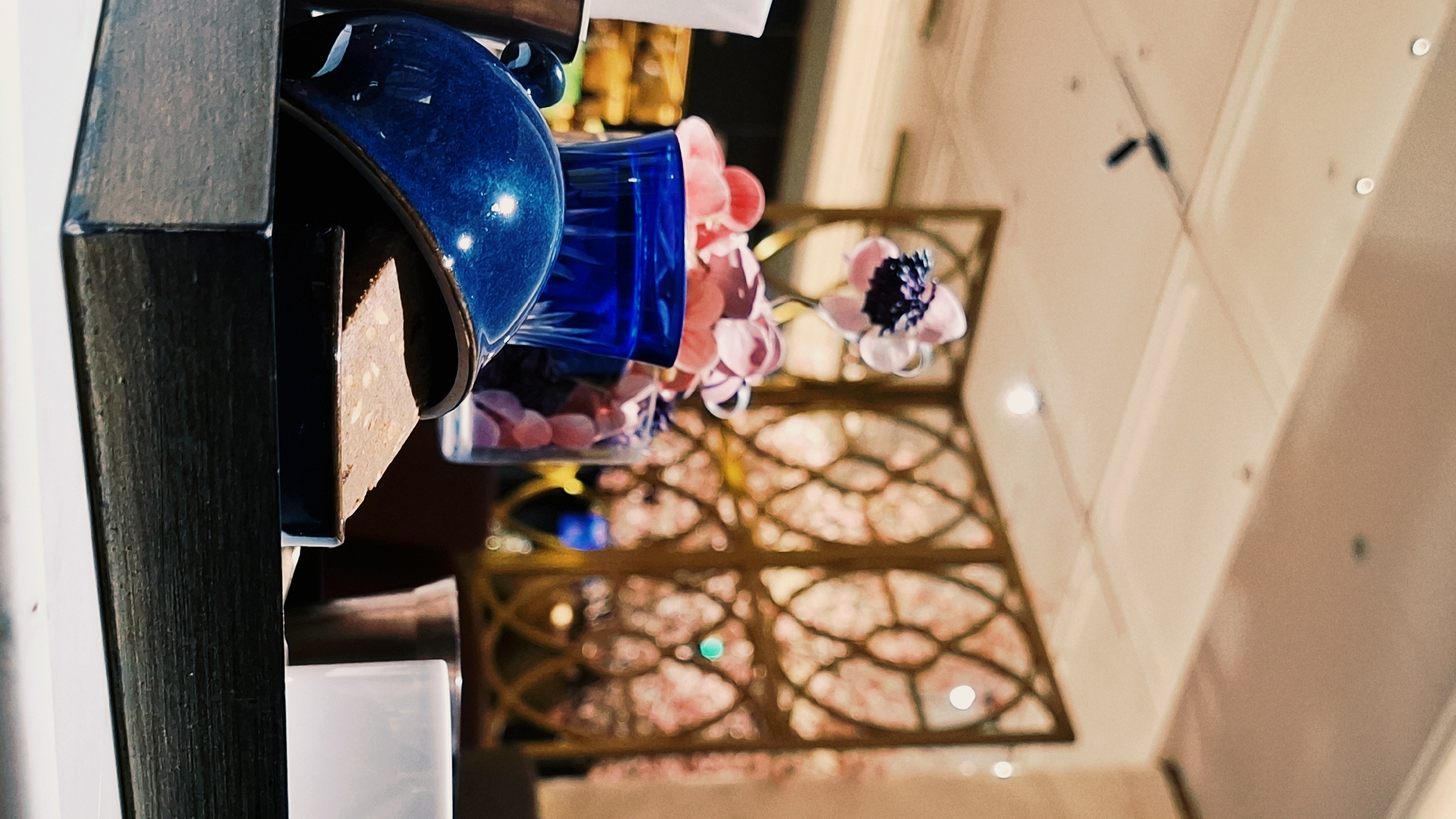 The image depicts an upward view from a table in an indoor setting. Objects on the table include a dark wood box, a blue ceramic container, a clear glass filled with pink items, and a floral arrangement with pink and white flowers. Overhead, there is a decorative gold-colored lattice design, and a light-colored ceiling. This image suggests a setting in Dongcheng District, China, potentially in a restaurant or interior design with local artistic expressions.